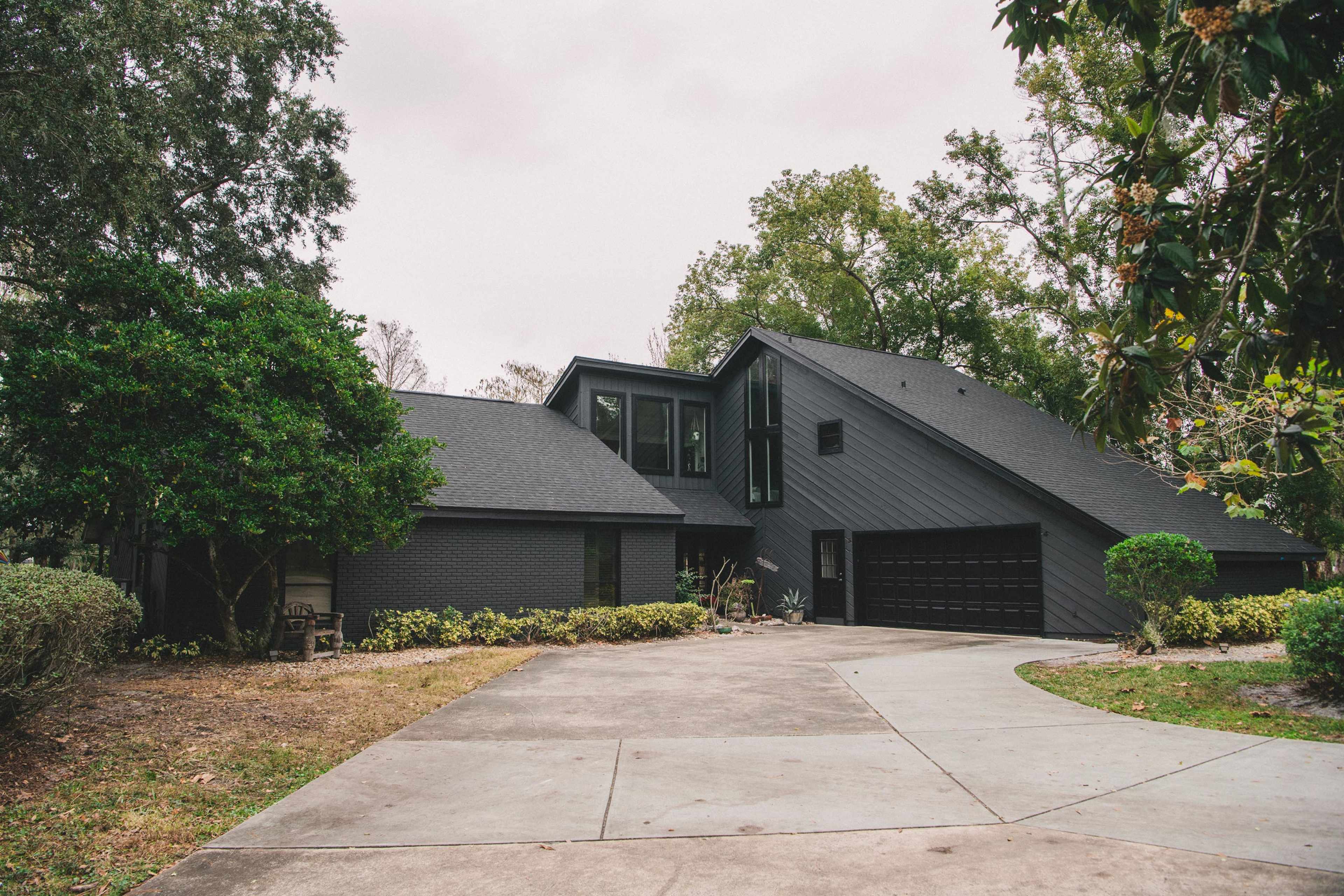 A modern black house with a sloped roof sits at the end of a curved driveway surrounded by greenery.