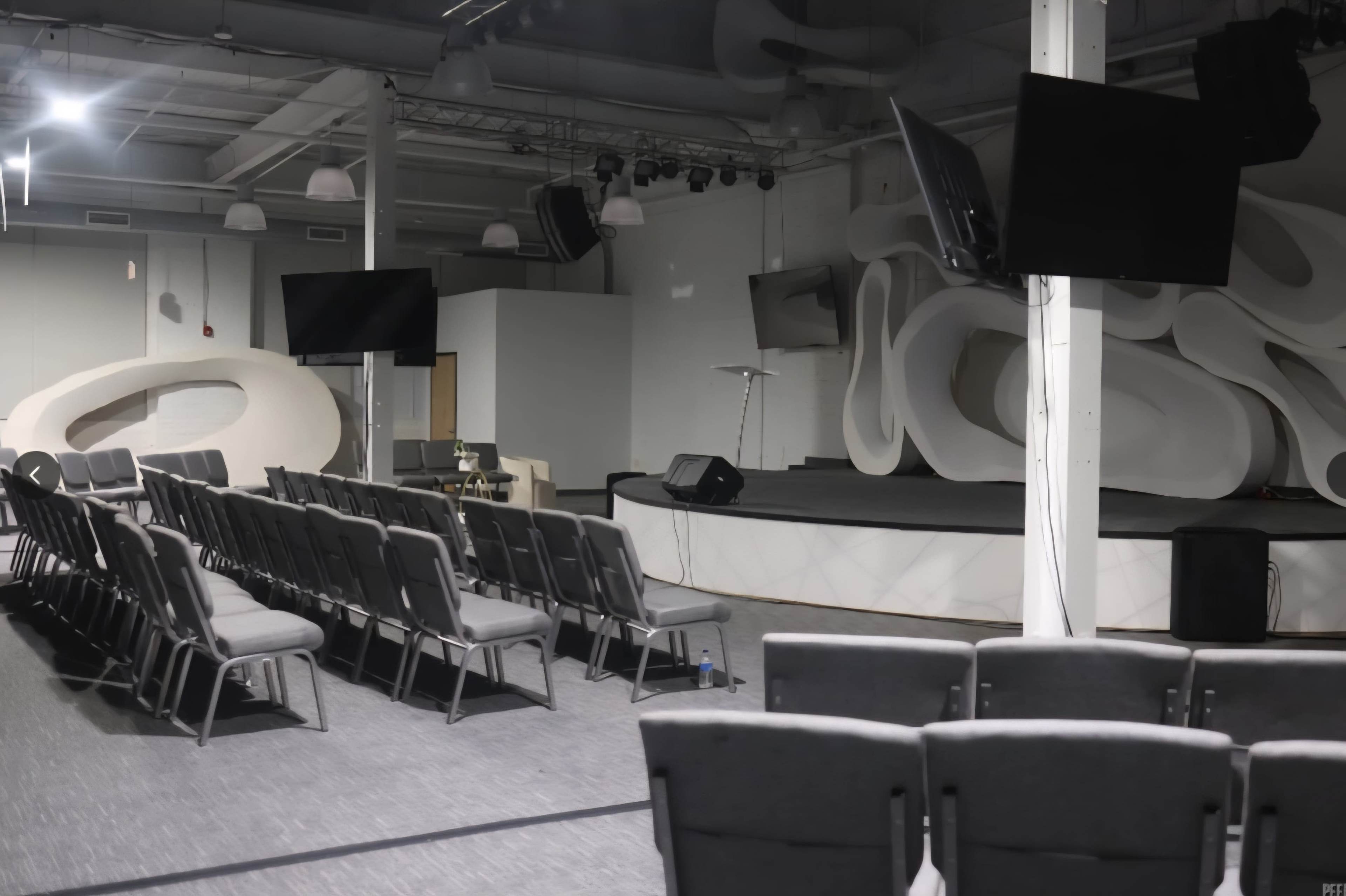 The image shows an interior space set up for a gathering, featuring rows of gray chairs facing a stage with a podium, surrounded by modern decor and screens.