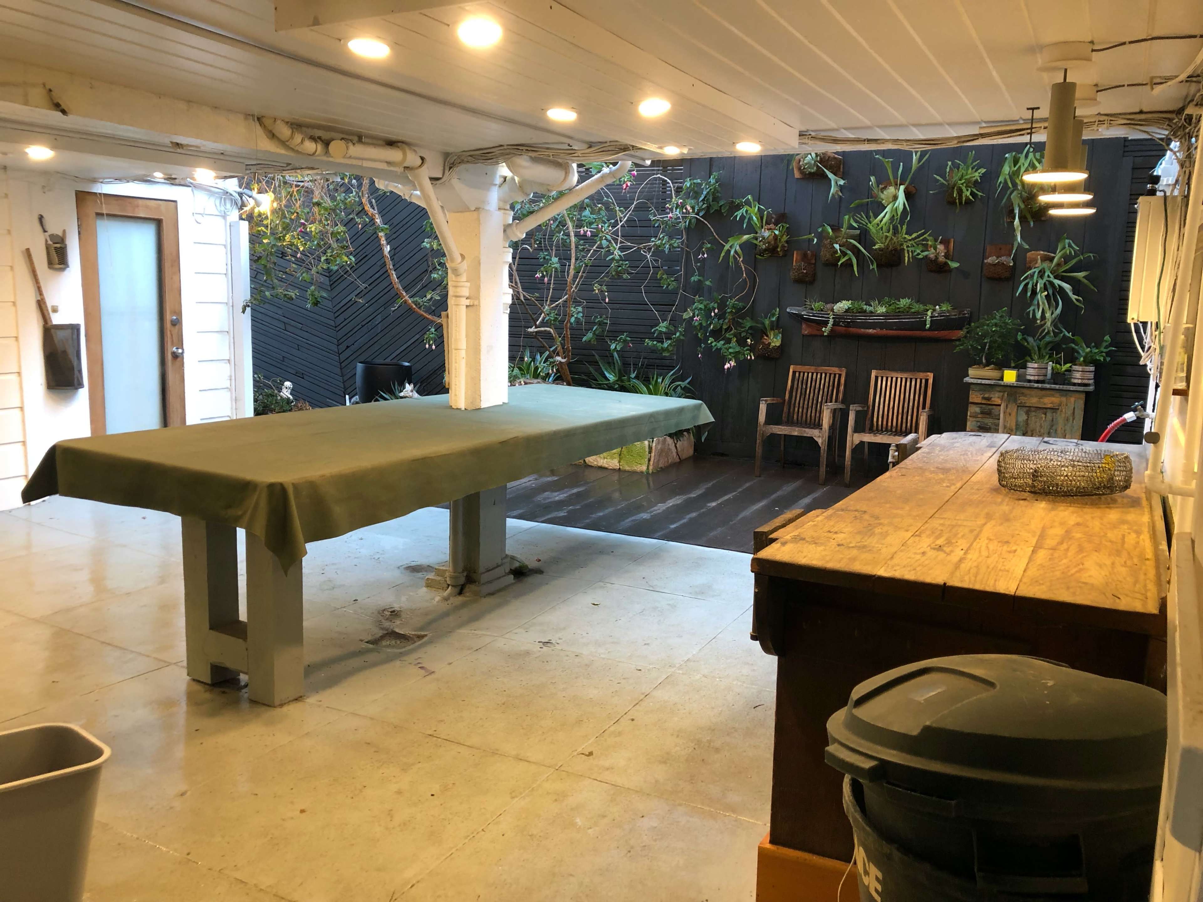 A spacious indoor area with a large green table, wooden furniture against a dark wall, and various plants hanging and arranged on the wall.