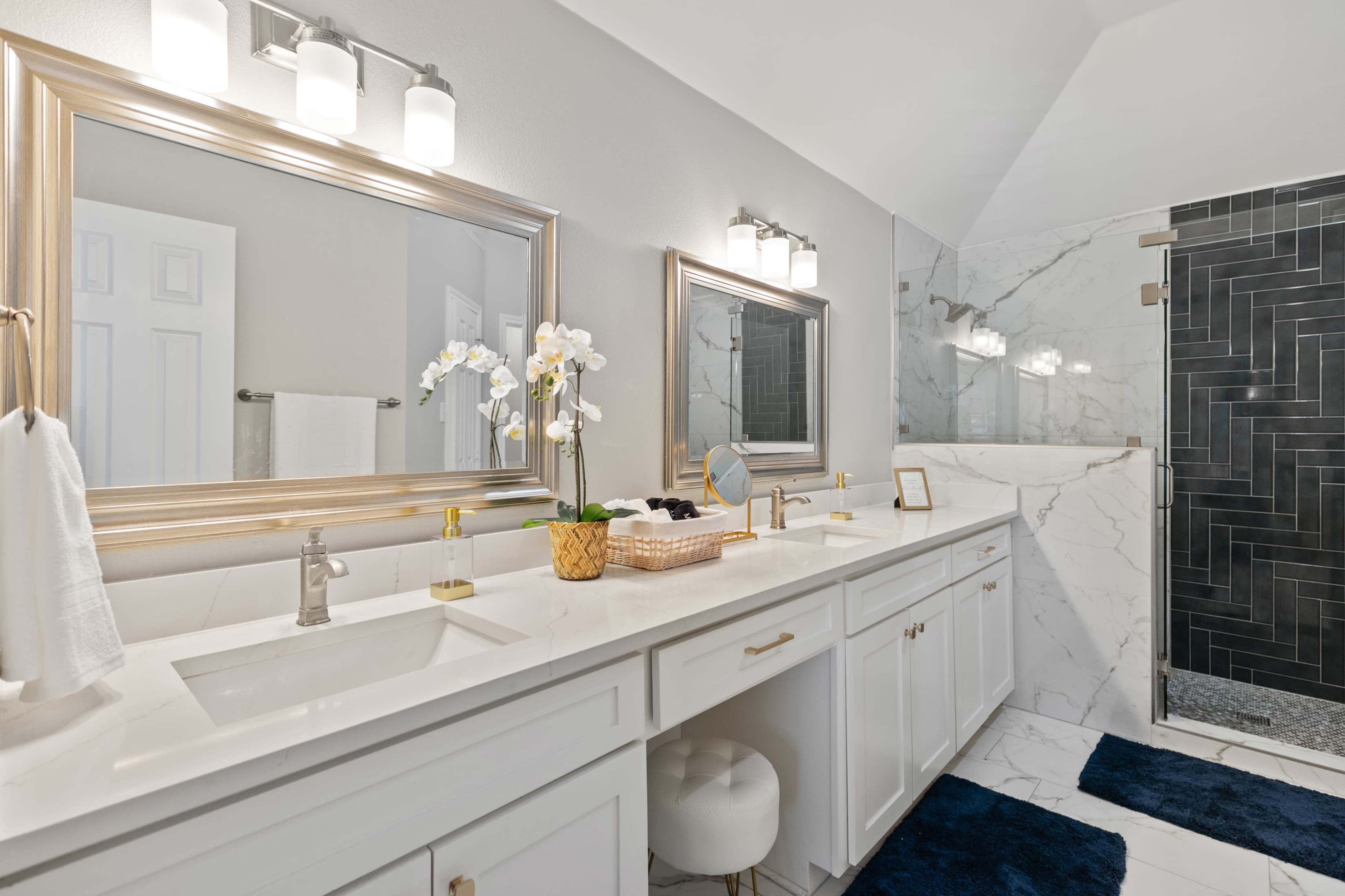 The image shows a modern bathroom with dual sinks, marble countertops, large mirrors framed in gold, and black tiled accents in the shower area.