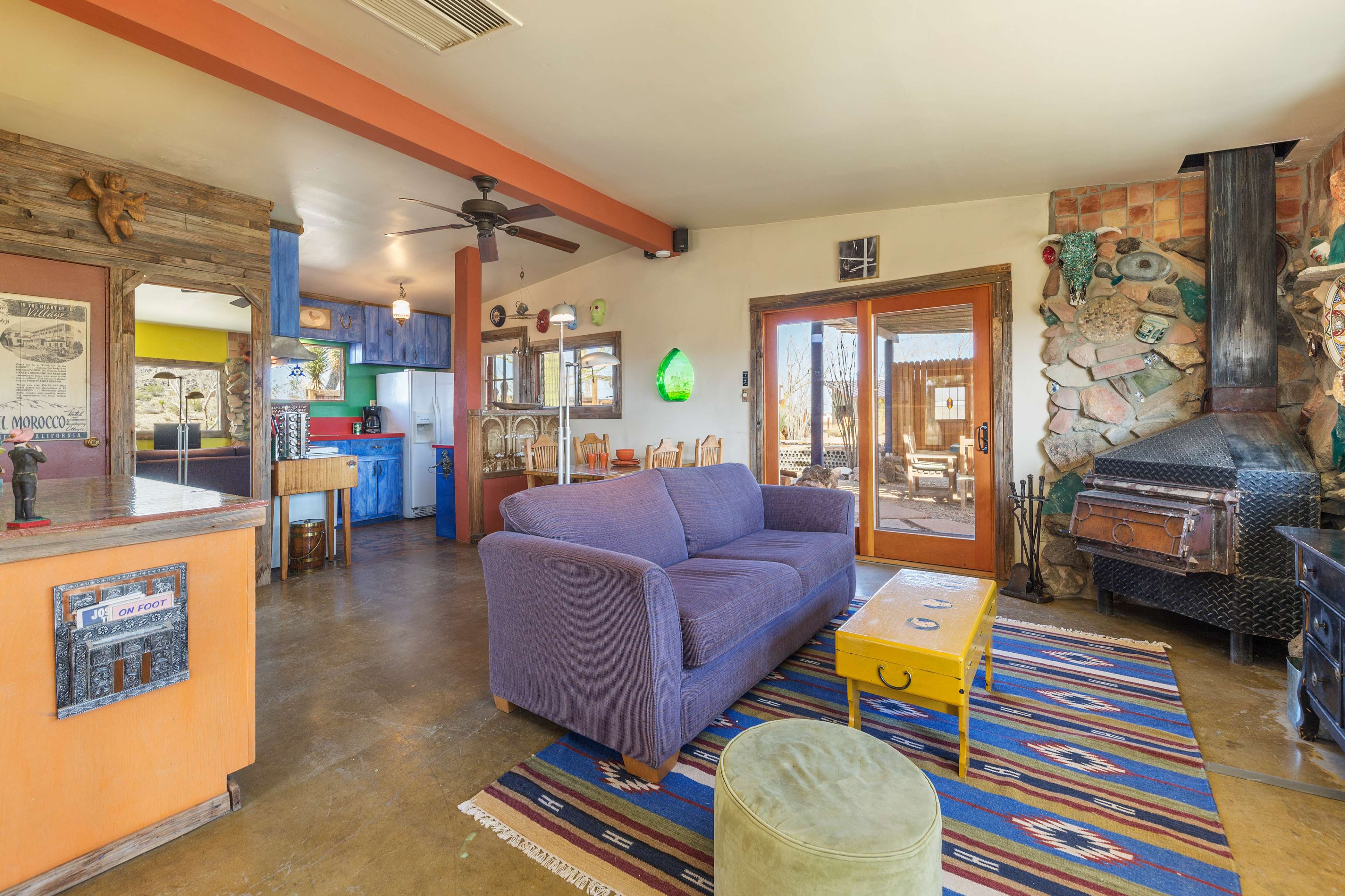 A cozy living room with a purple sofa, a yellow coffee table, and a stone fireplace, featuring an open layout that connects to a kitchen area.