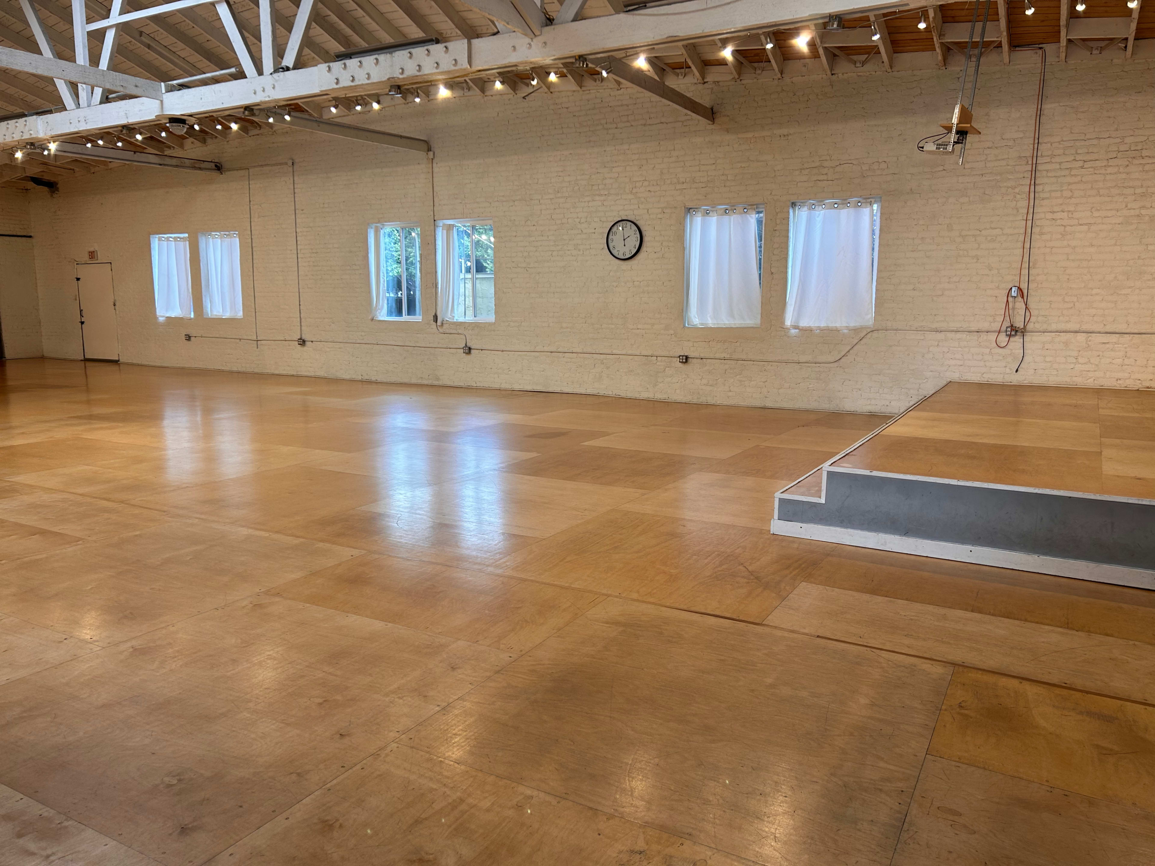 A spacious room with wooden flooring, white walls, large windows, a clock on the wall, and a raised platform at one end.