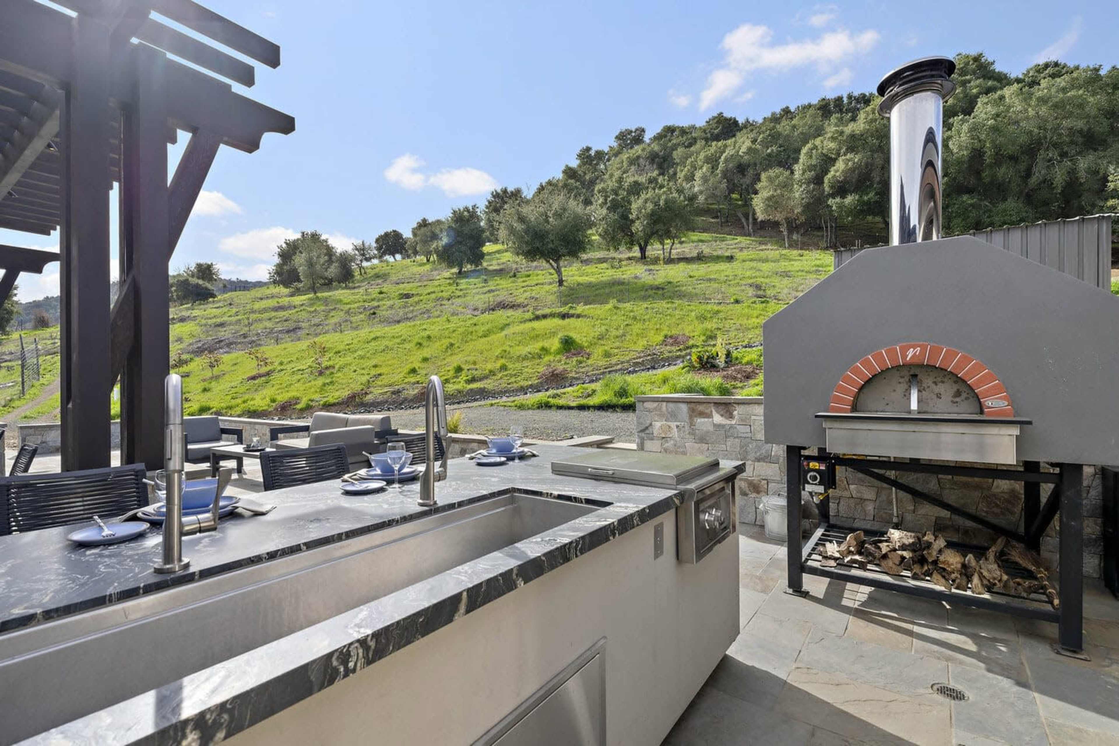 The image shows an outdoor kitchen area featuring a stone pizza oven and a stainless steel countertop with seating and table settings, set against a hillside with lush greenery.