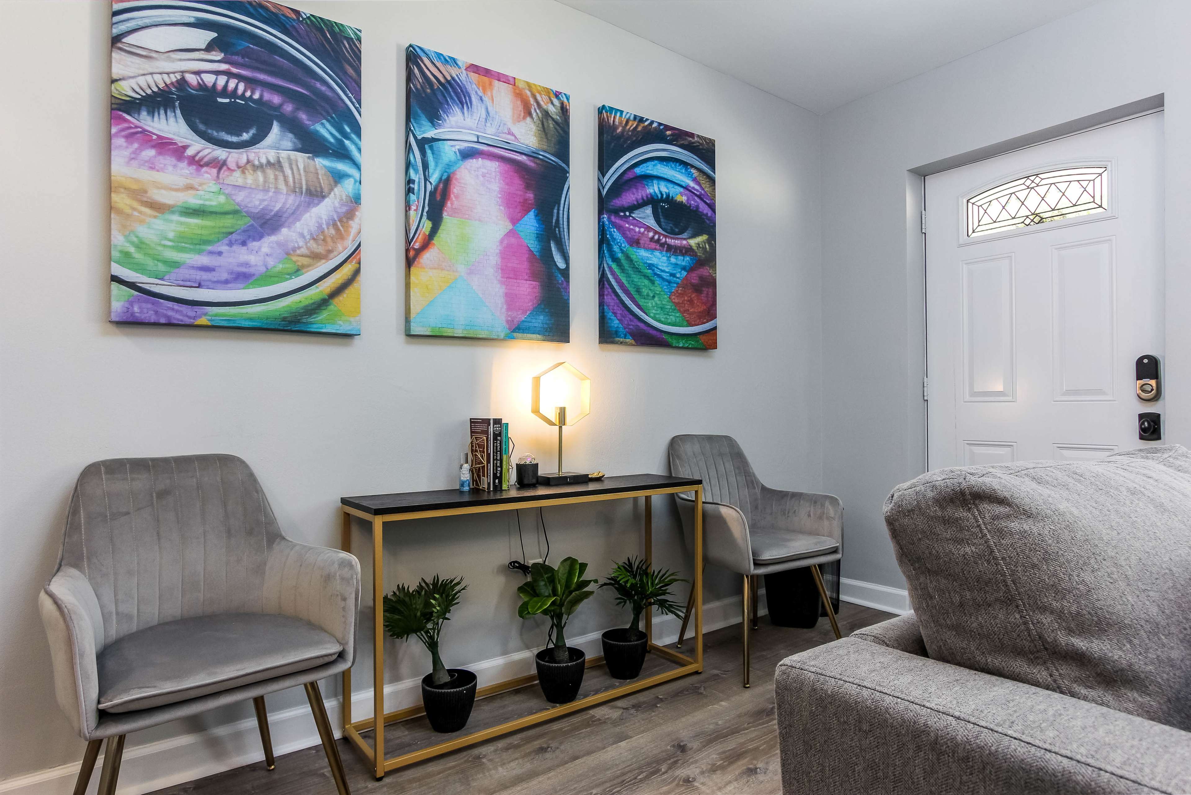 The image shows a modern living space featuring two gray chairs, a small table with a lamp, and three colorful eye-themed artworks on the wall, alongside a white front door.