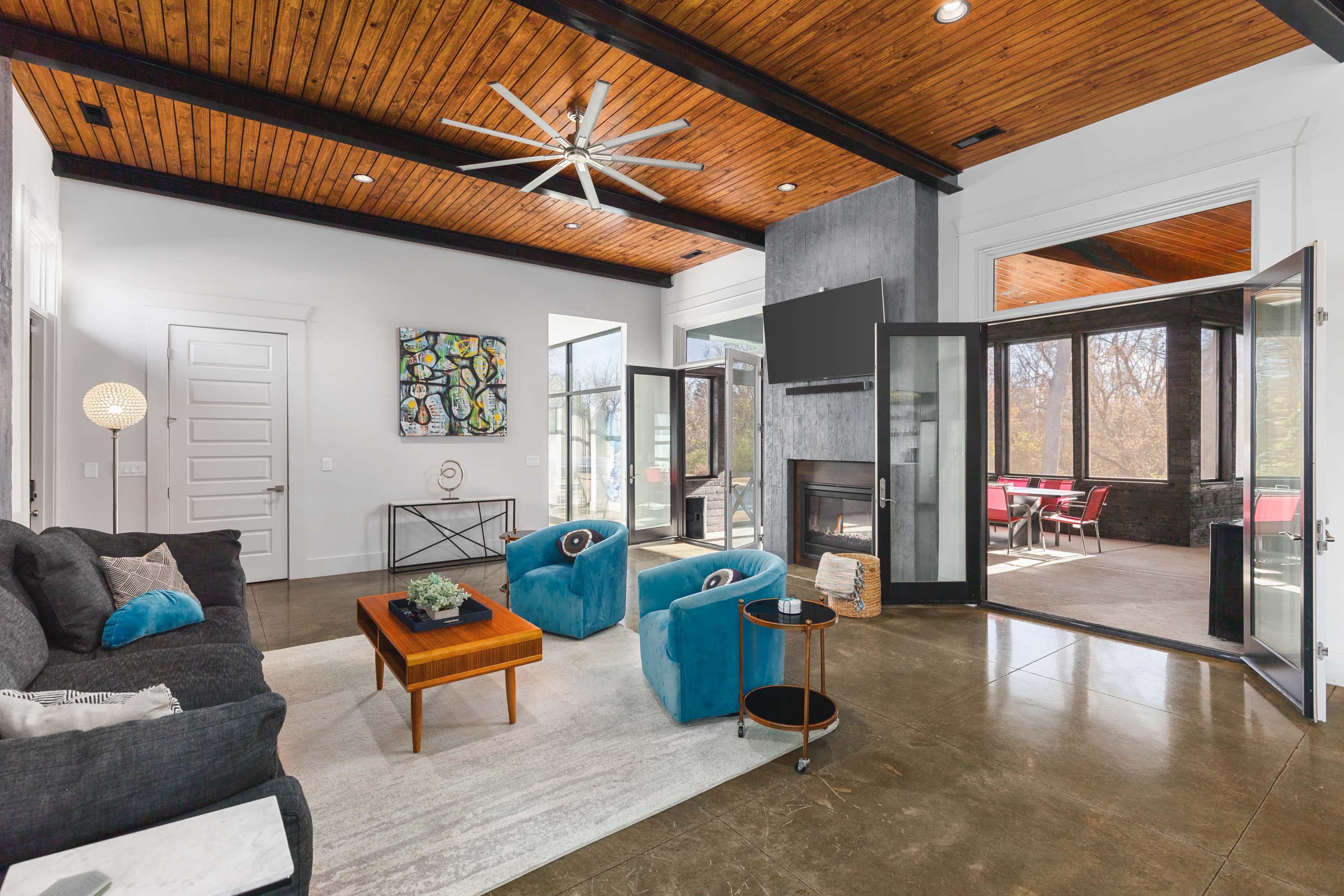 A modern living room with a dark couch, vibrant blue chairs, a wooden ceiling, and large glass doors leading to an outdoor area.