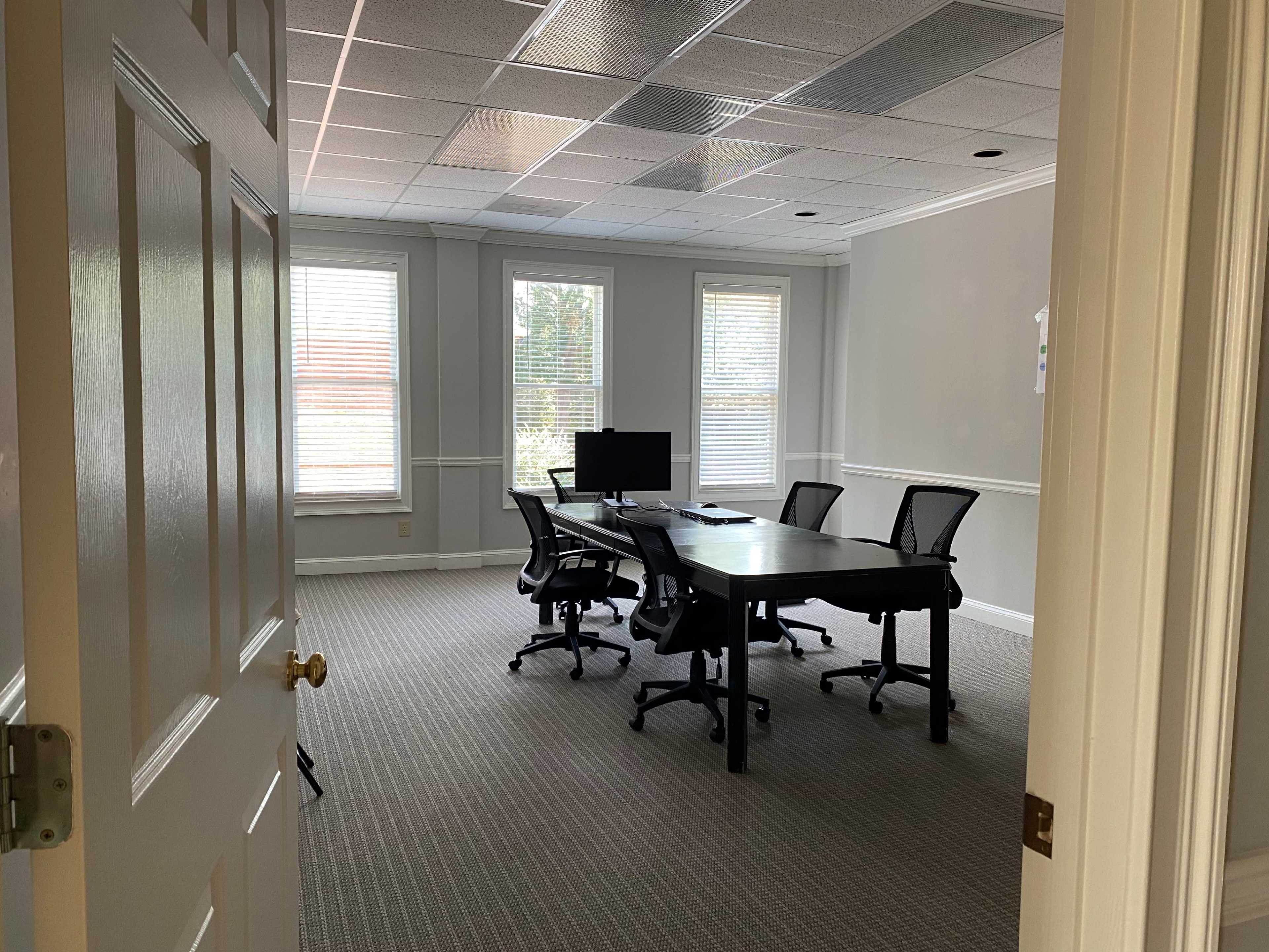 A vacant conference room with a large table, four chairs, and a computer monitor is visible through an open door.