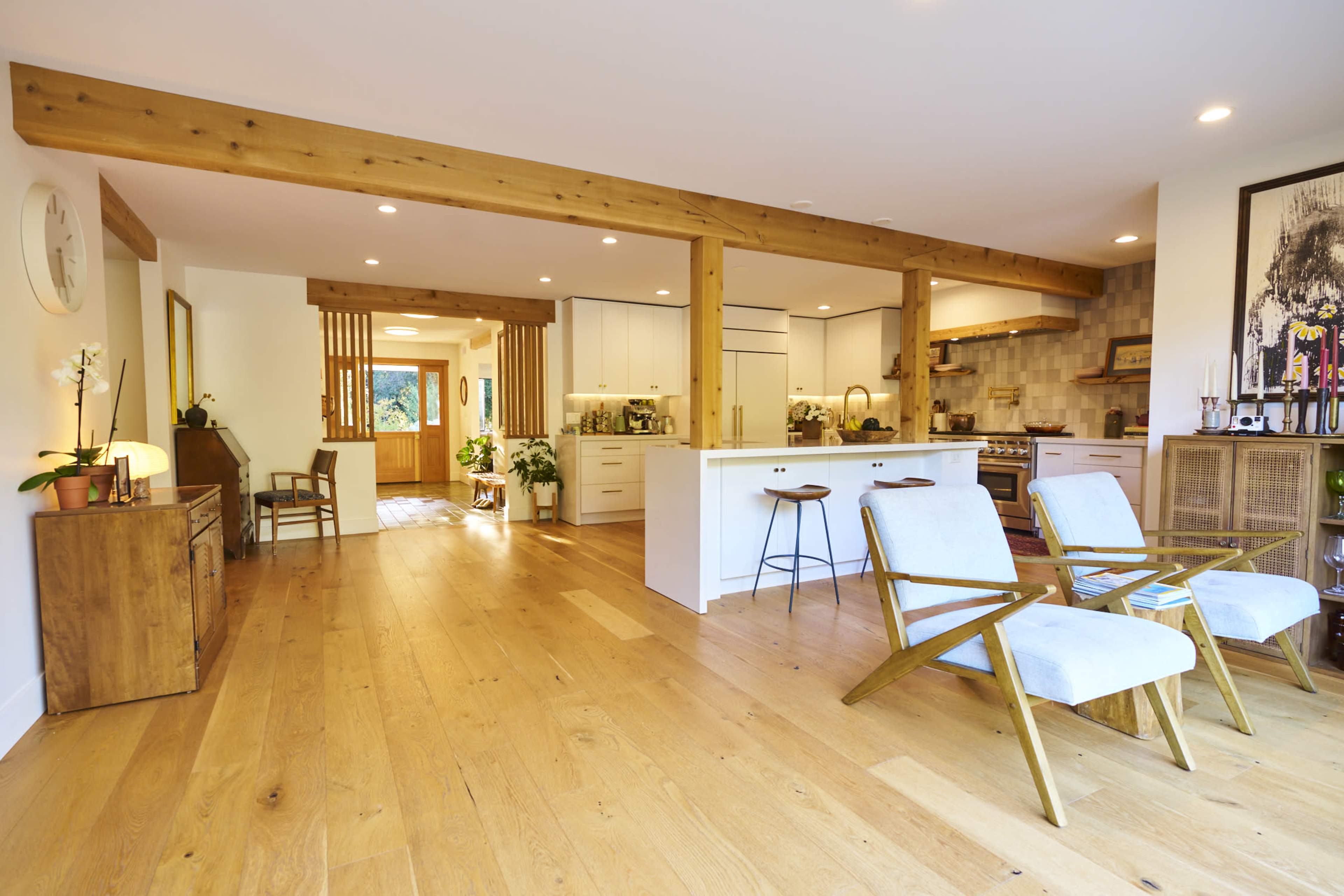 The image shows a modern open-concept living space featuring wooden beams, light-colored cabinetry, and two armchairs facing a kitchen area.