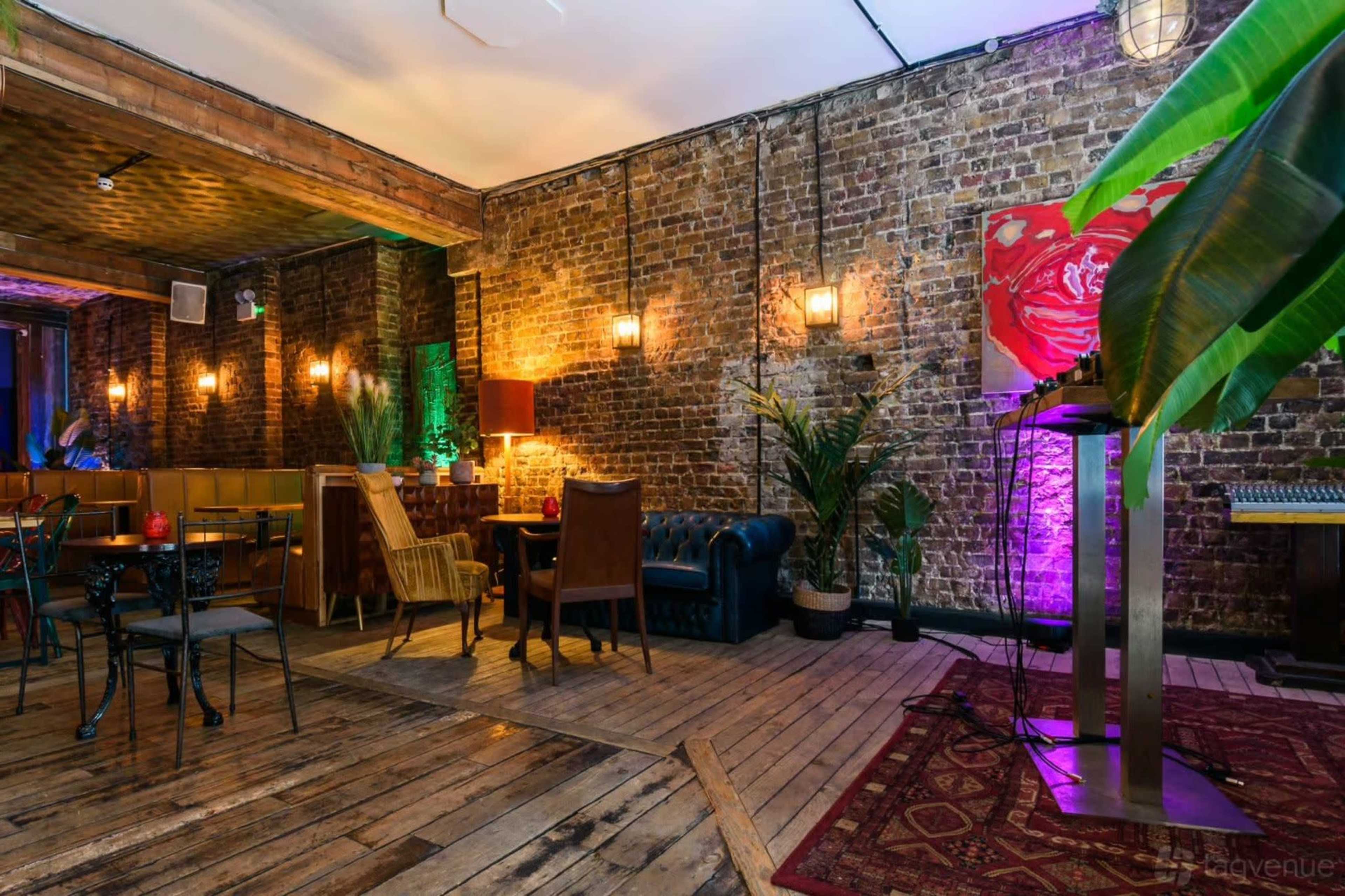 The image shows a stylish interior of a venue featuring exposed brick walls, wooden floors, and various seating arrangements with plants and decorative lighting.