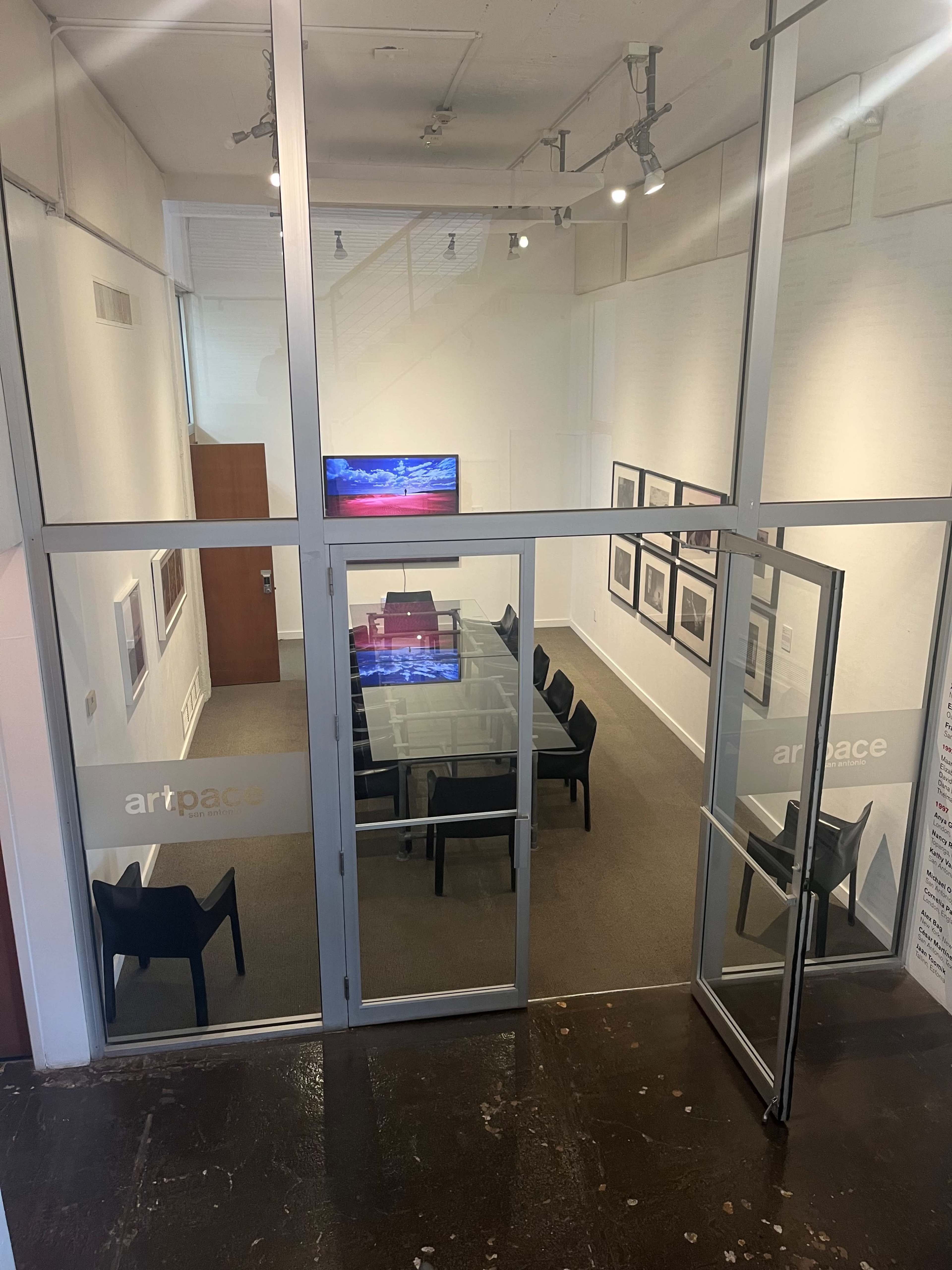 The image shows a conference room with a glass wall, a large table surrounded by black chairs, and an empty display screen in front of a white wall with framed photos.