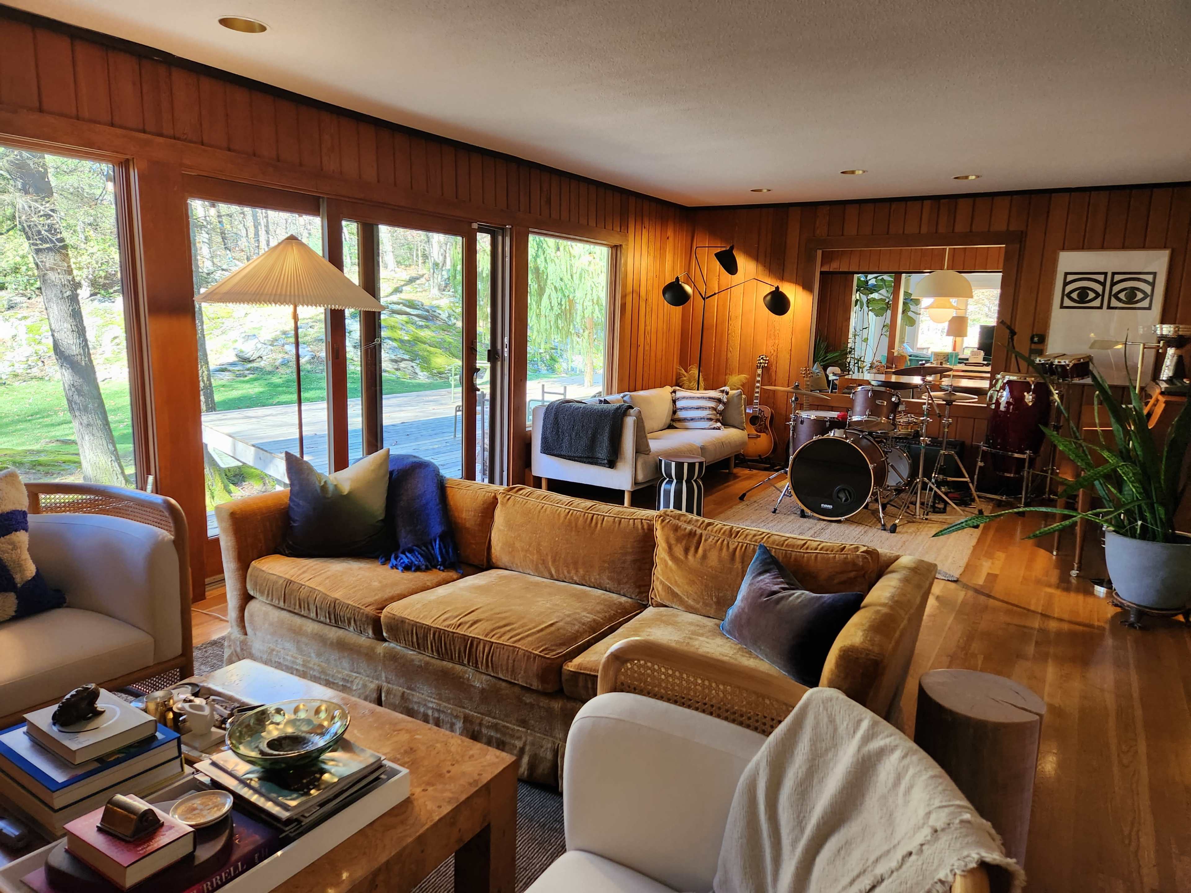 The image shows a cozy living room with a brown sofa, a small dining area in the background, and large windows that reveal a view of greenery outside.