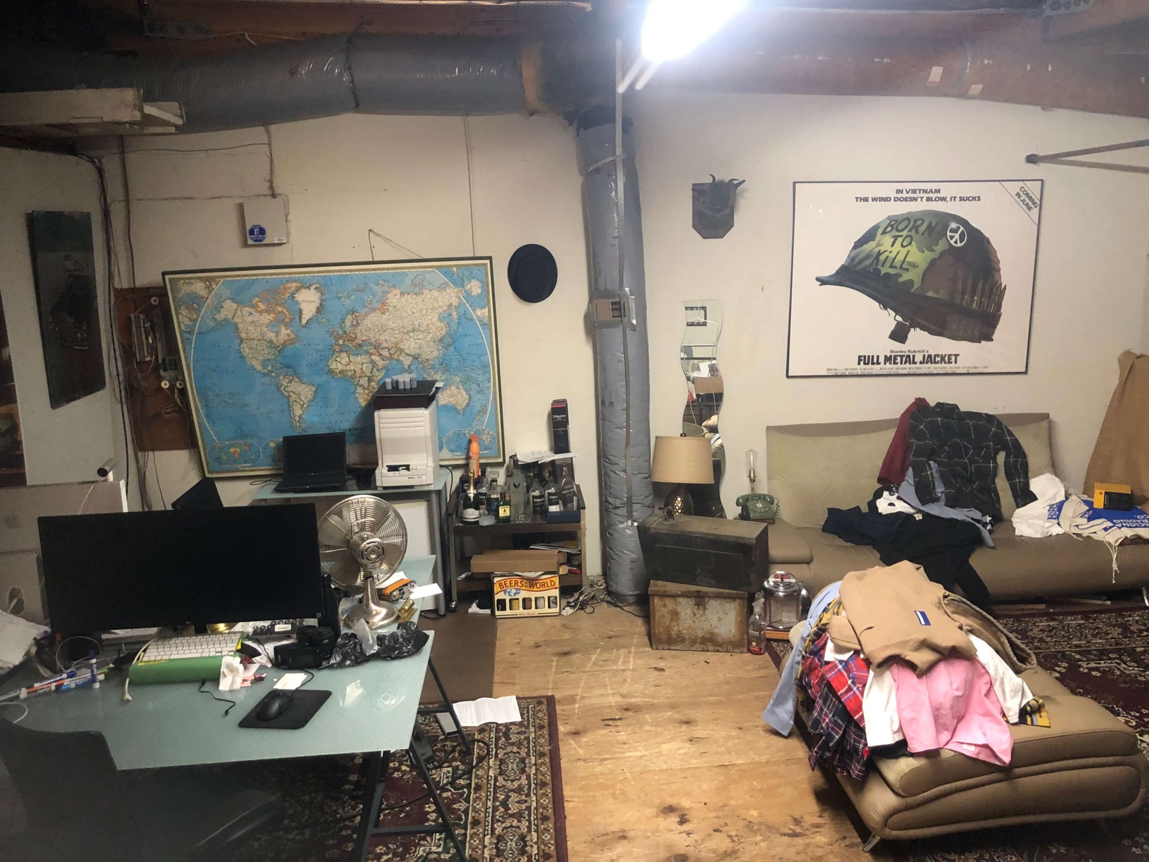 A cluttered room with a map on the wall, a computer setup on a desk, and various items scattered on a sofa and the floor.