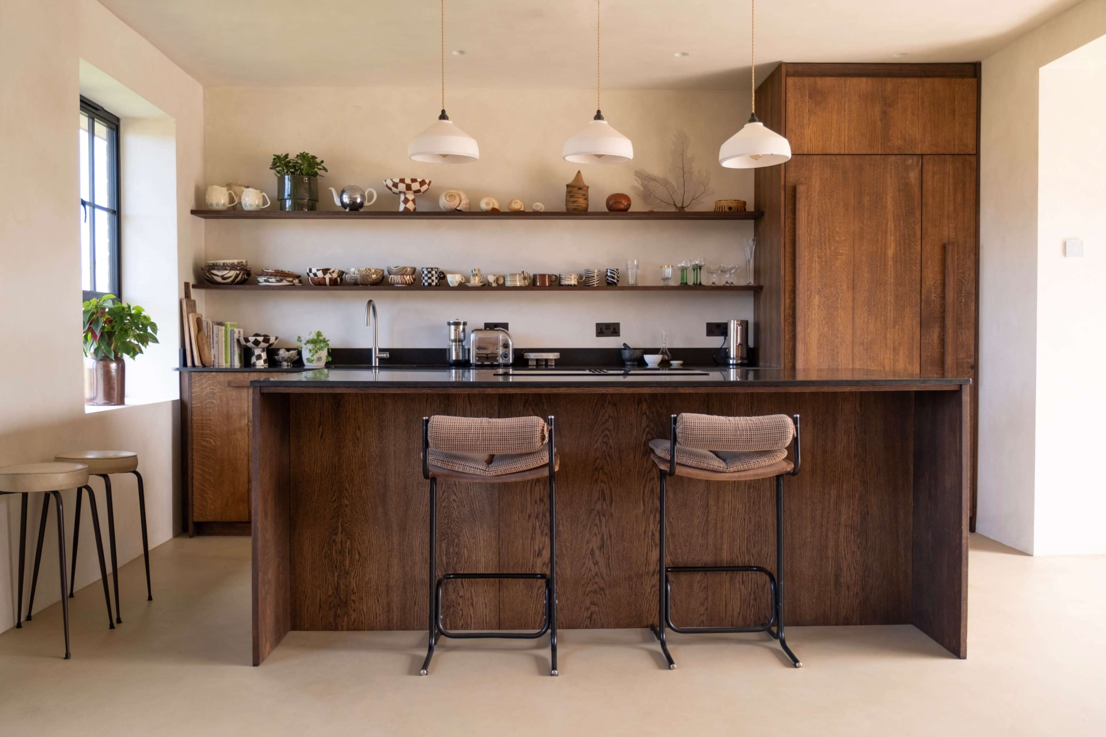 The image shows a modern kitchen with wooden cabinetry, a central island featuring two bar stools, and shelves displaying various kitchen items.