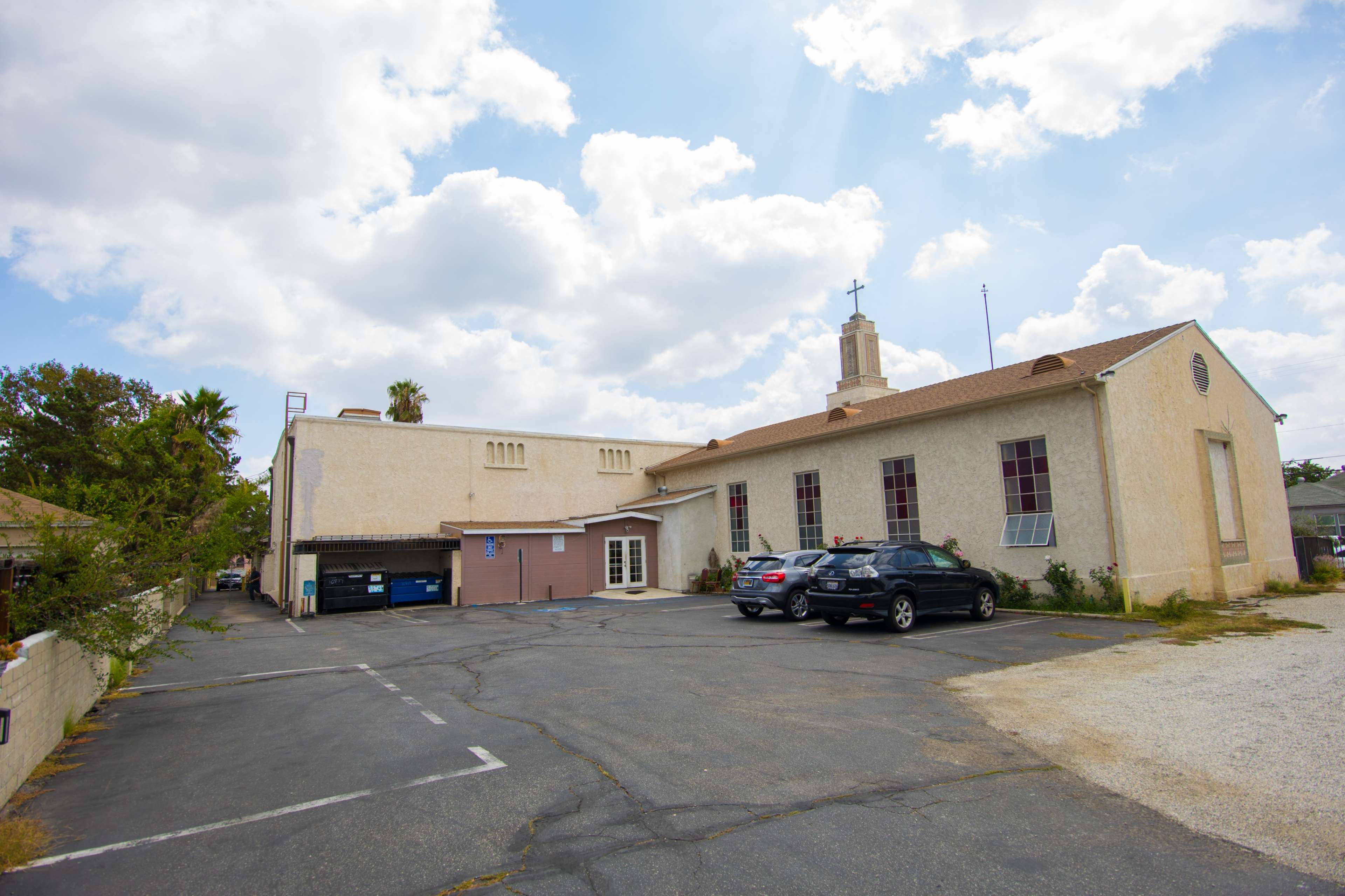A light-colored building with a steeple and cross is situated next to a parking area that contains several vehicles, including SUVs, with a few trees visible nearby.