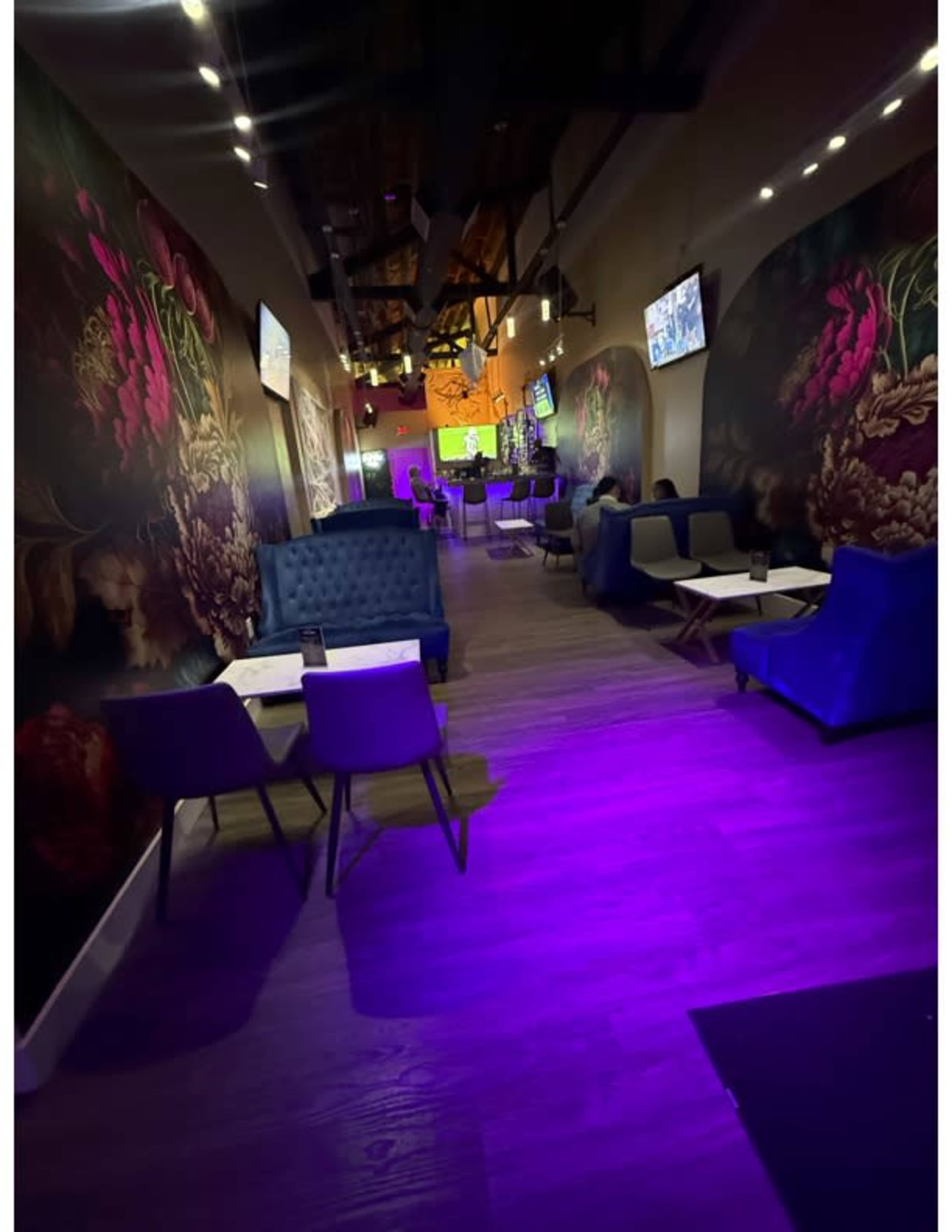 The image shows a modern lounge area with decorative floral wall art, dim lighting, and a mix of seating arrangements including blue velvet chairs and small tables.