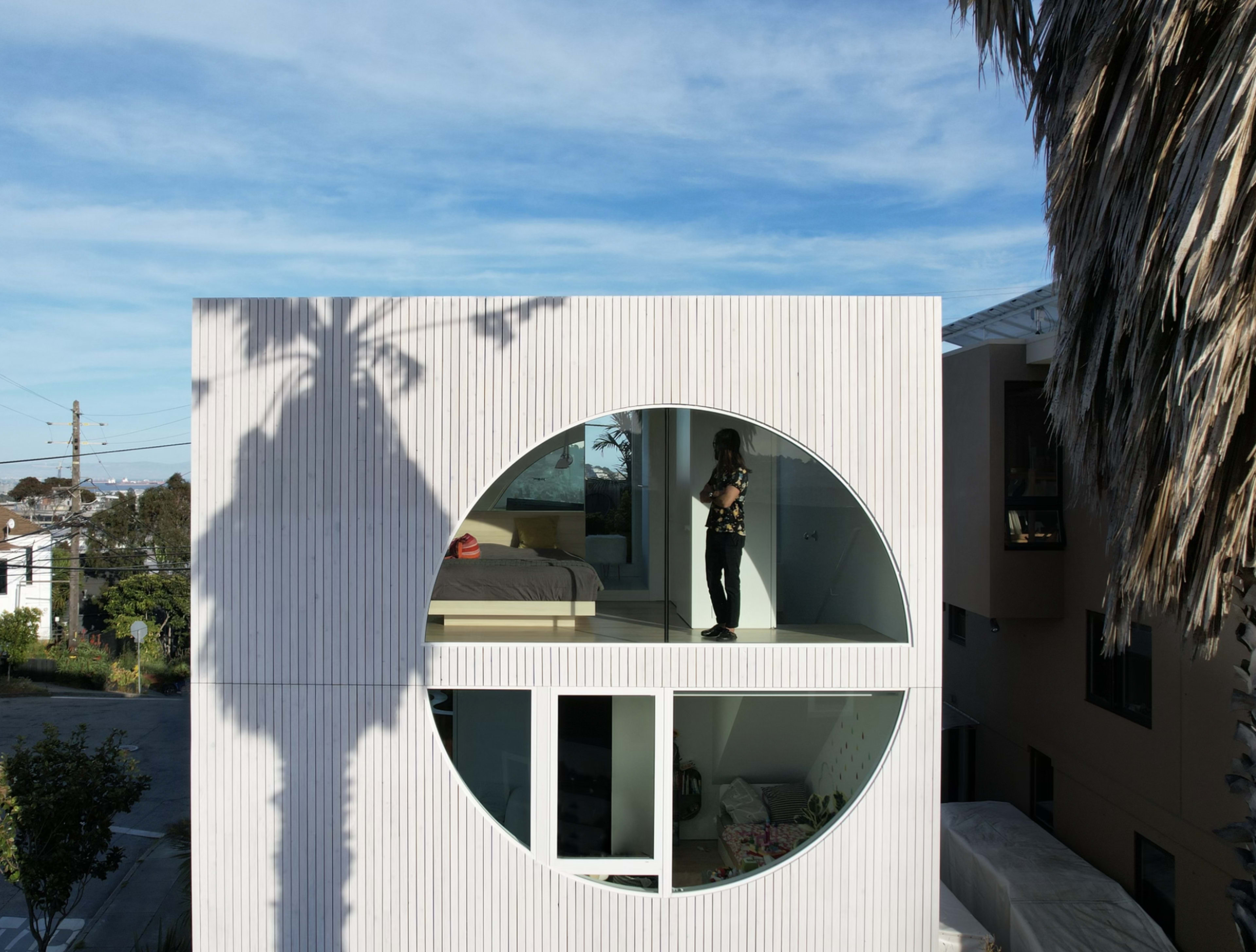 A modern building with a large circular window features a person standing inside, while the shadow of a nearby palm tree is cast on the exterior wall.