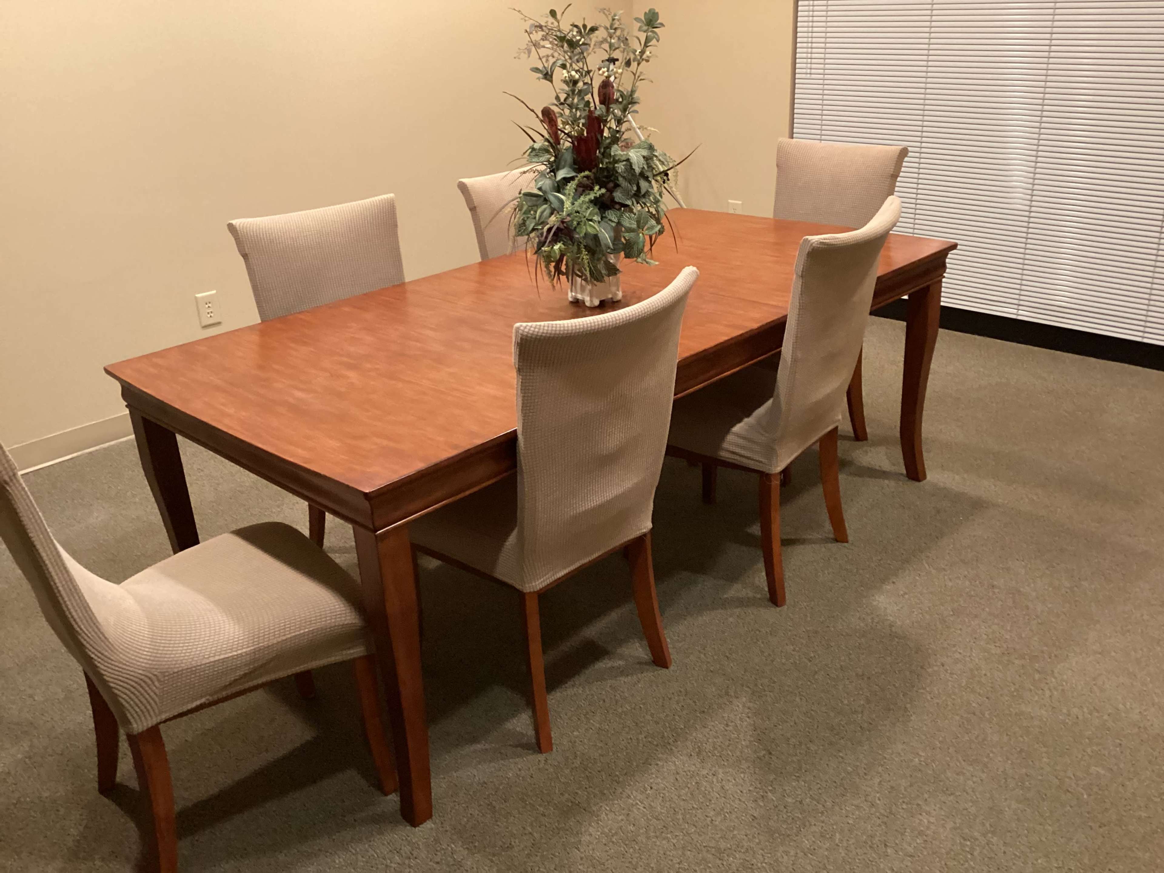 A wooden dining table with six upholstered chairs and a decorative plant arrangement sits in a room with carpeted flooring.