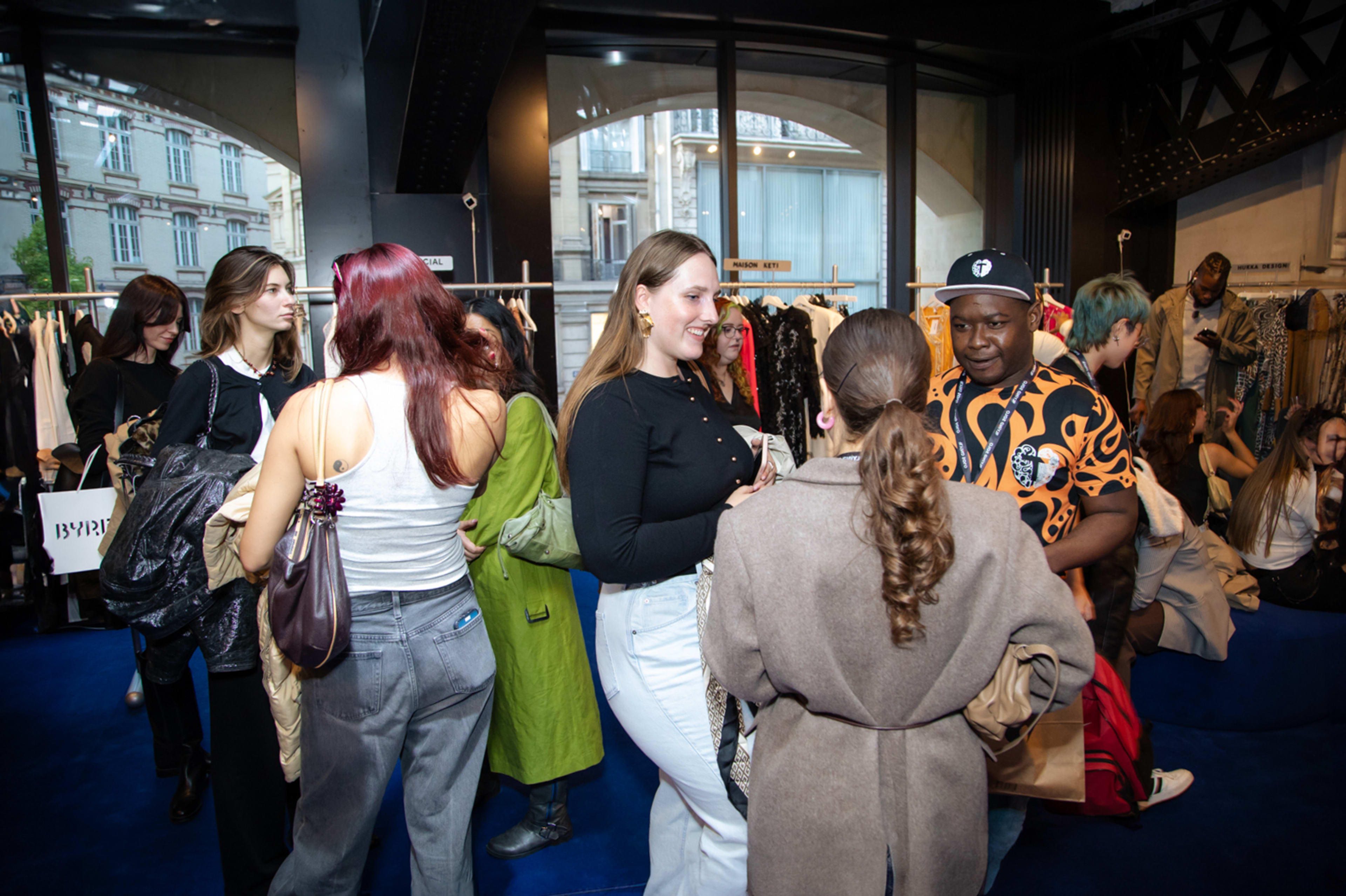 A group of people socialize and browse clothing in a vibrant retail space with large windows.