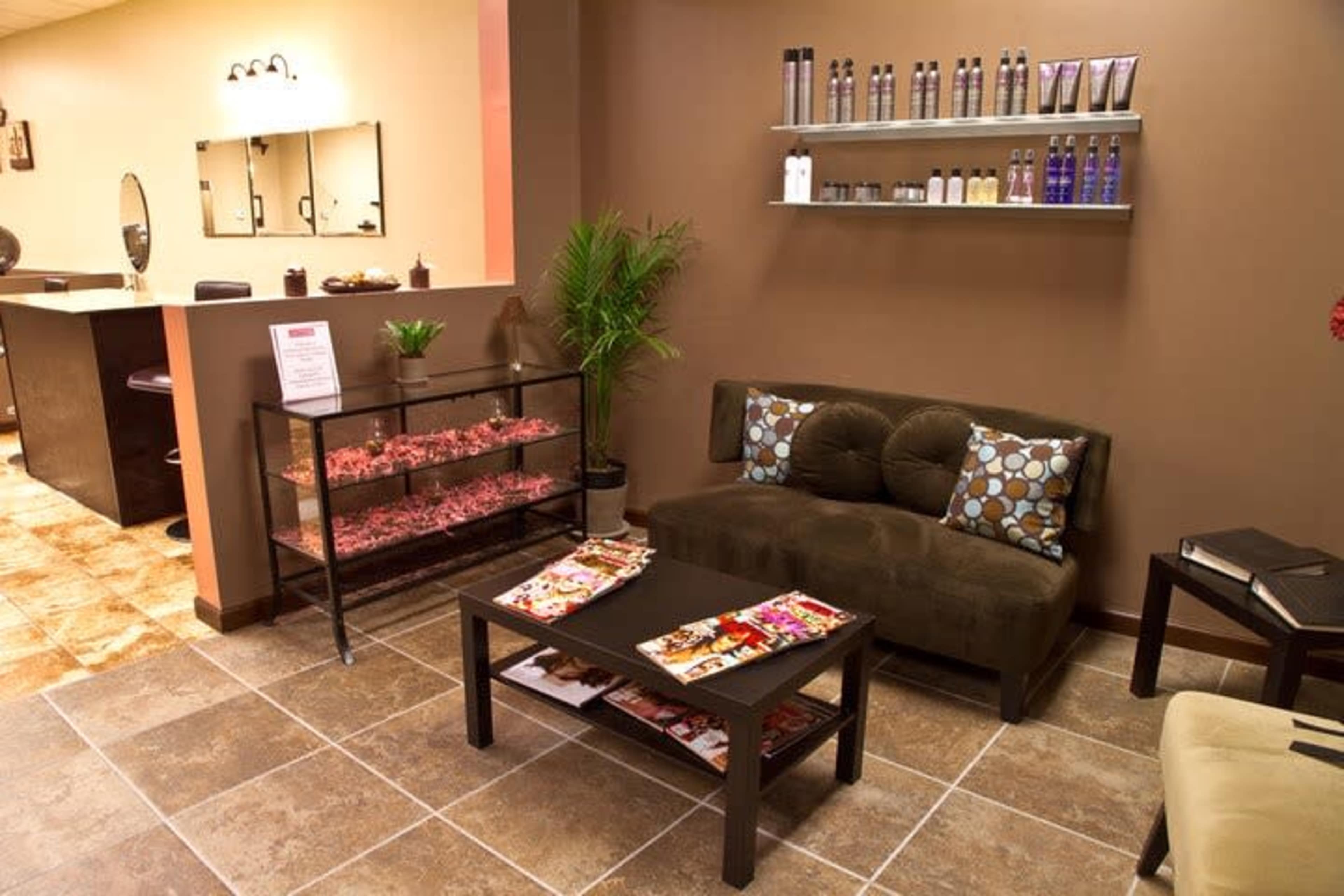 The image shows a salon waiting area featuring a brown couch with patterned cushions, a coffee table with magazines, and a glass display case filled with products, all set against a muted wall color.