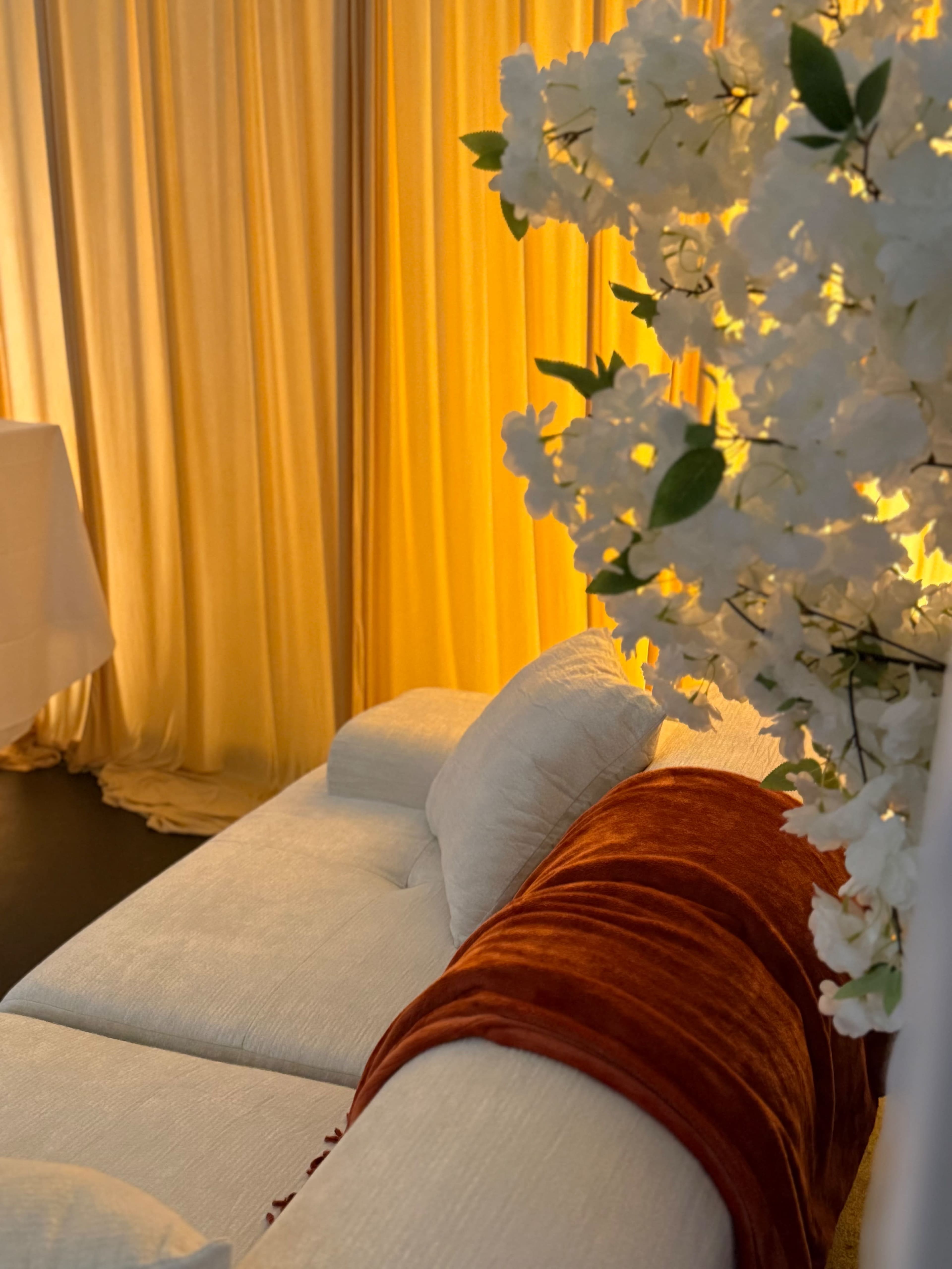 A white sofa with an orange throw is positioned next to a display of white flowers, set against warm yellow curtains.