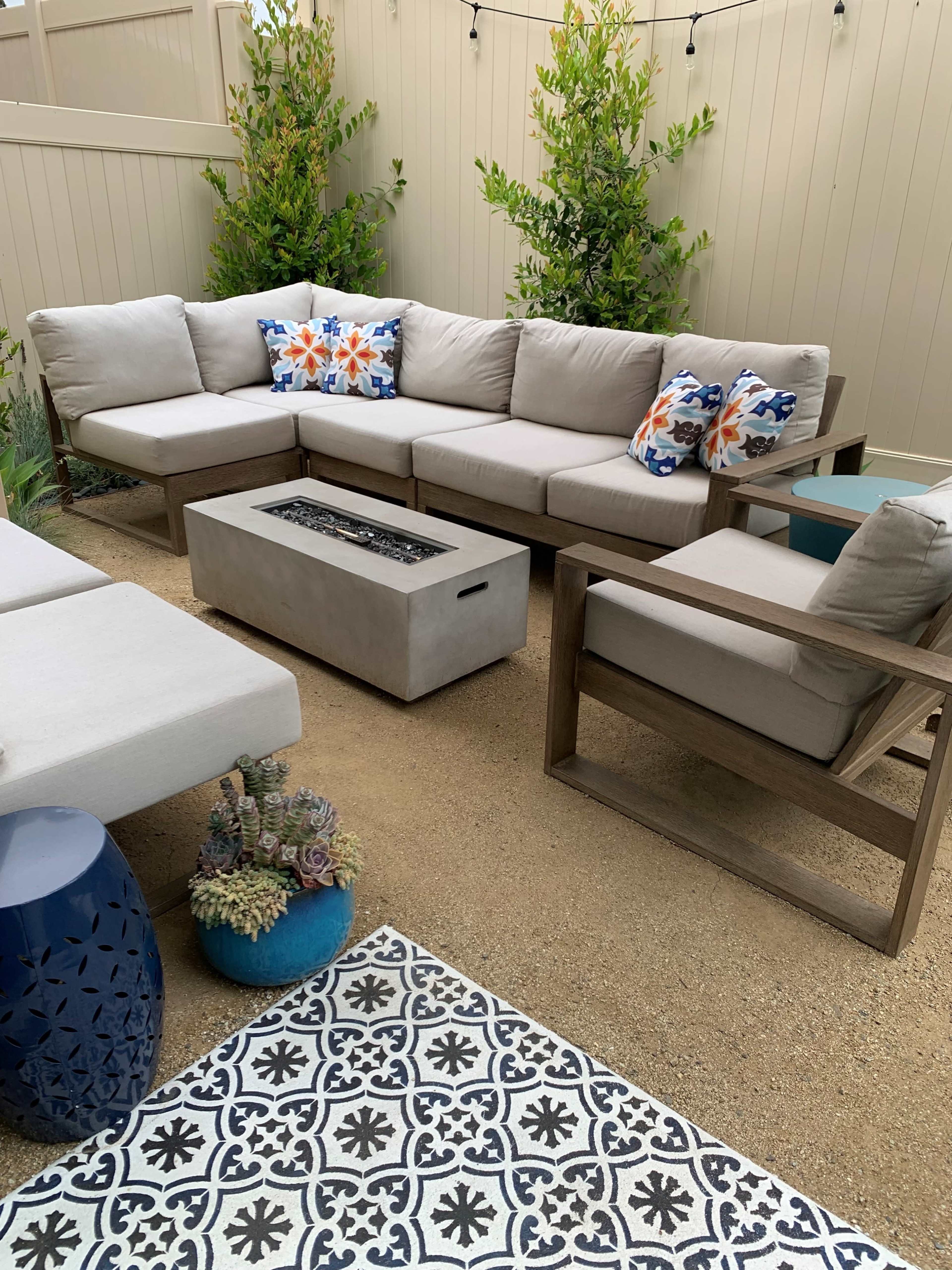 The image shows a modern outdoor seating area with a sectional sofa, a fire pit table, two armchairs, and decorative pillows on a patterned rug.