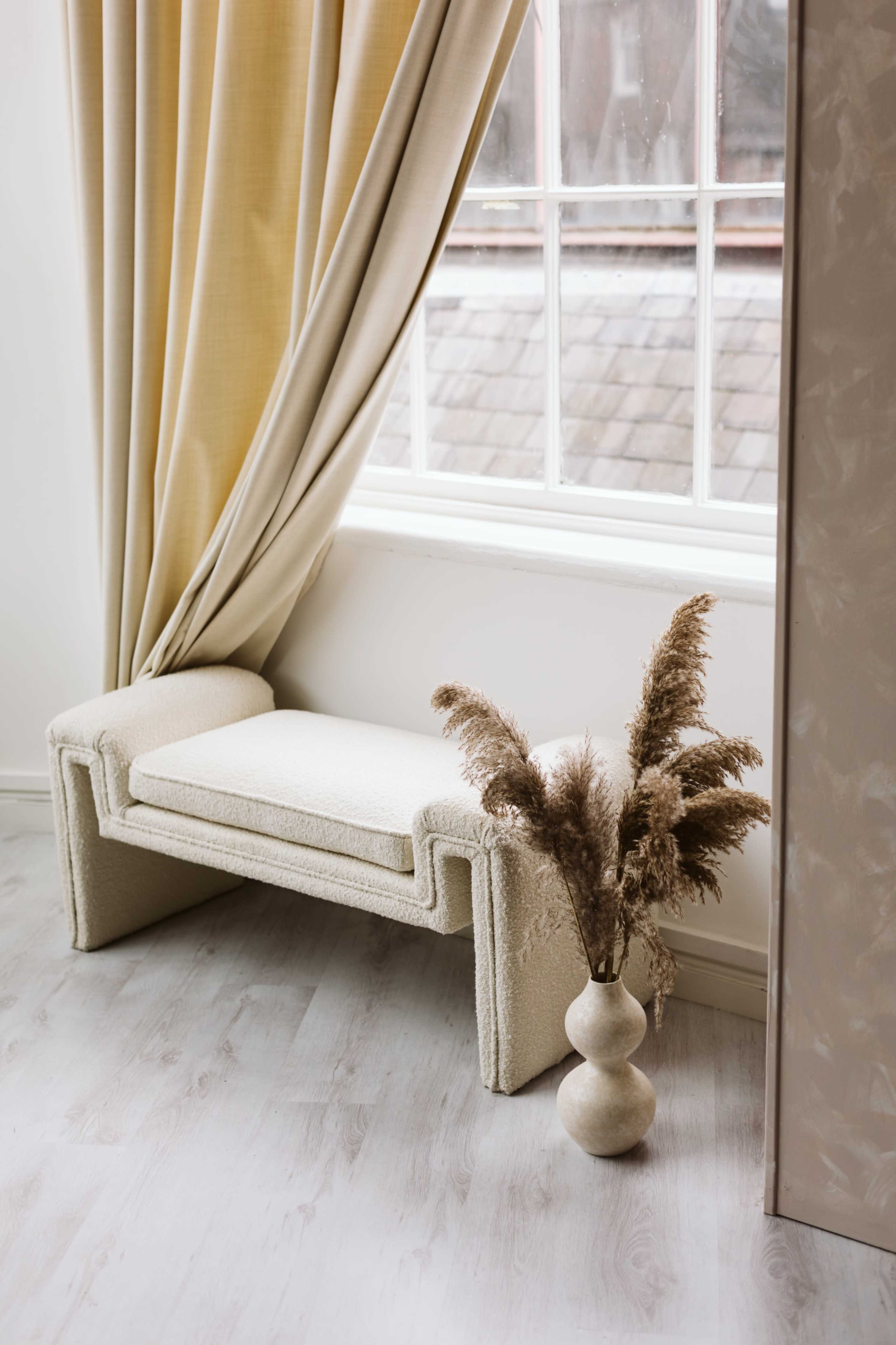 A textured bench with a rounded base and a vase holding dried pampas grass is positioned near a window with light-colored curtains.