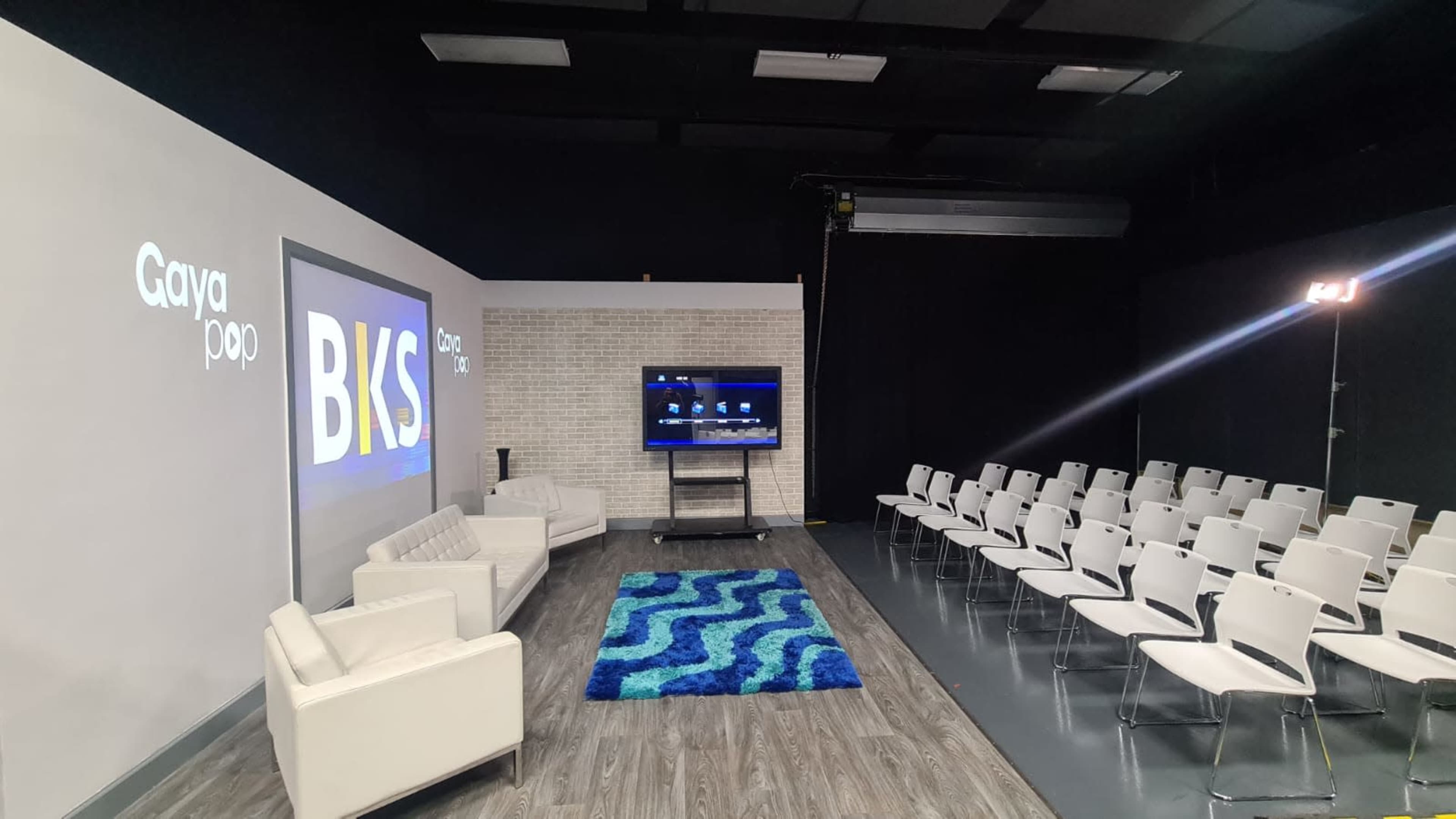 The image shows a studio setting with white walls, a blue carpet, a television on a stand, and rows of white chairs facing the screen.