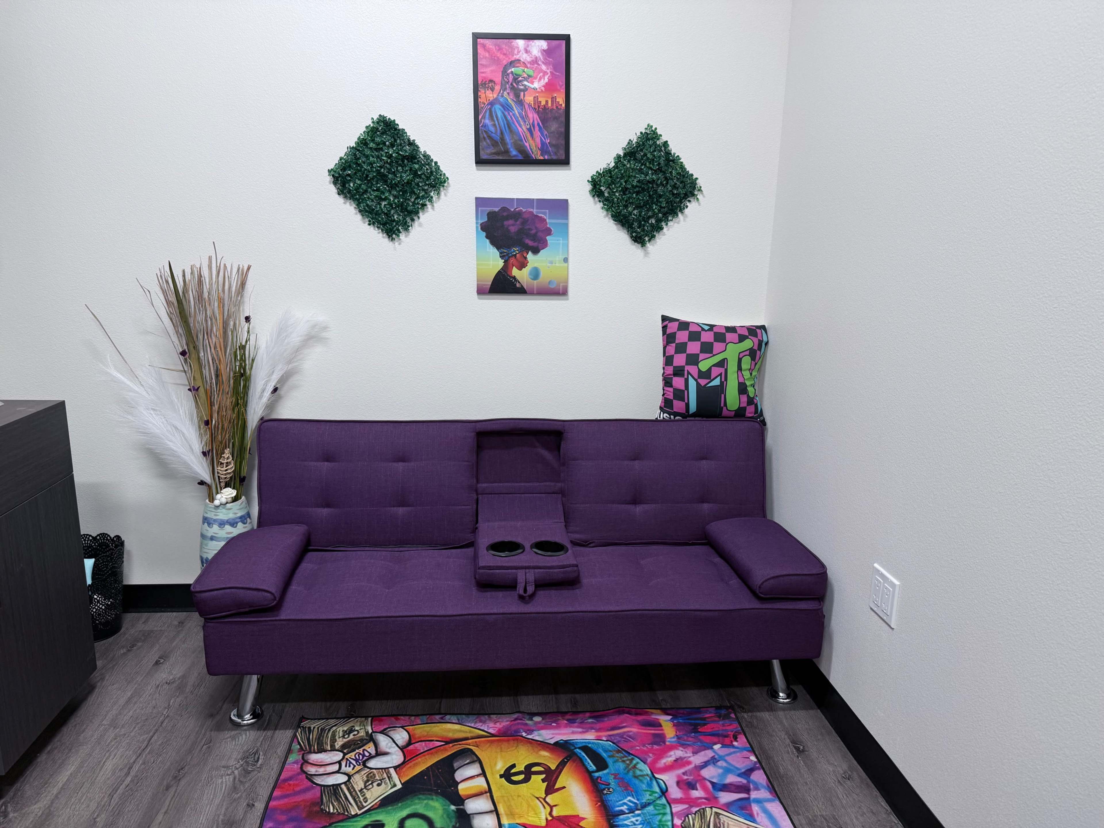 The image shows a purple sofa positioned against a light-colored wall, decorated with four wall hangings and a colorful rug on the floor.