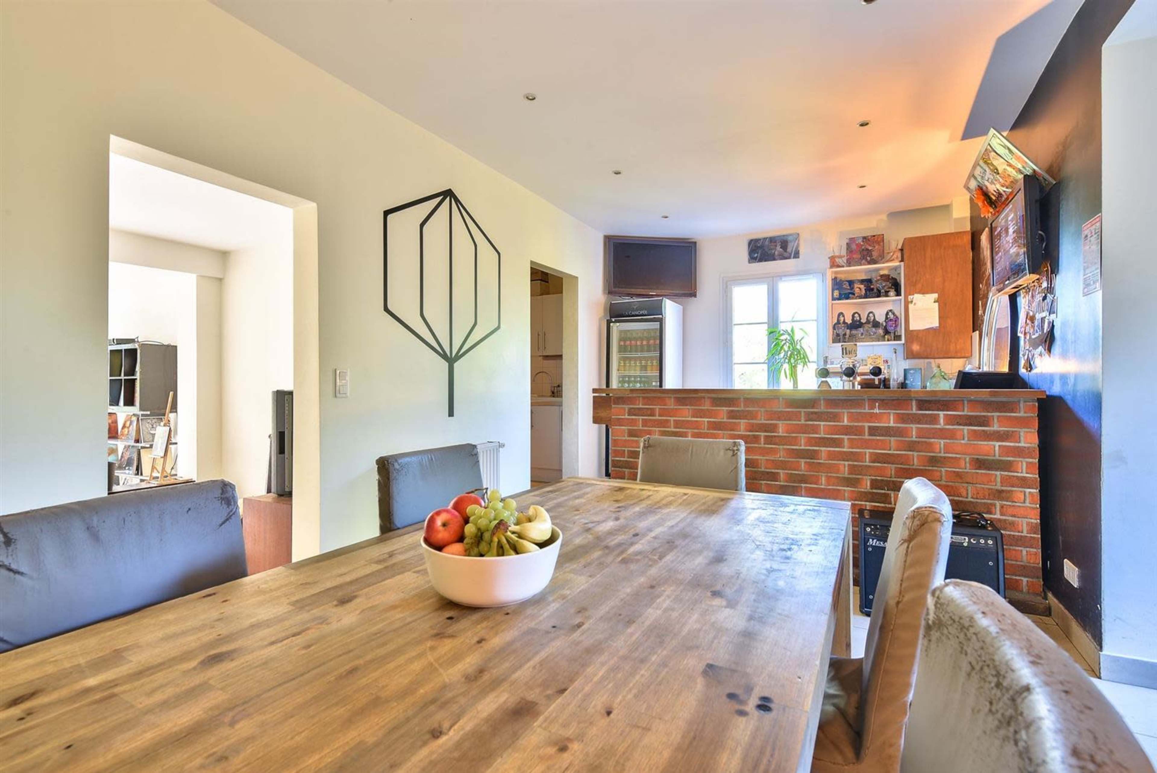 A wooden dining table with a bowl of fruit is set in a kitchen area featuring a brick wall, cabinets, and various decorations.