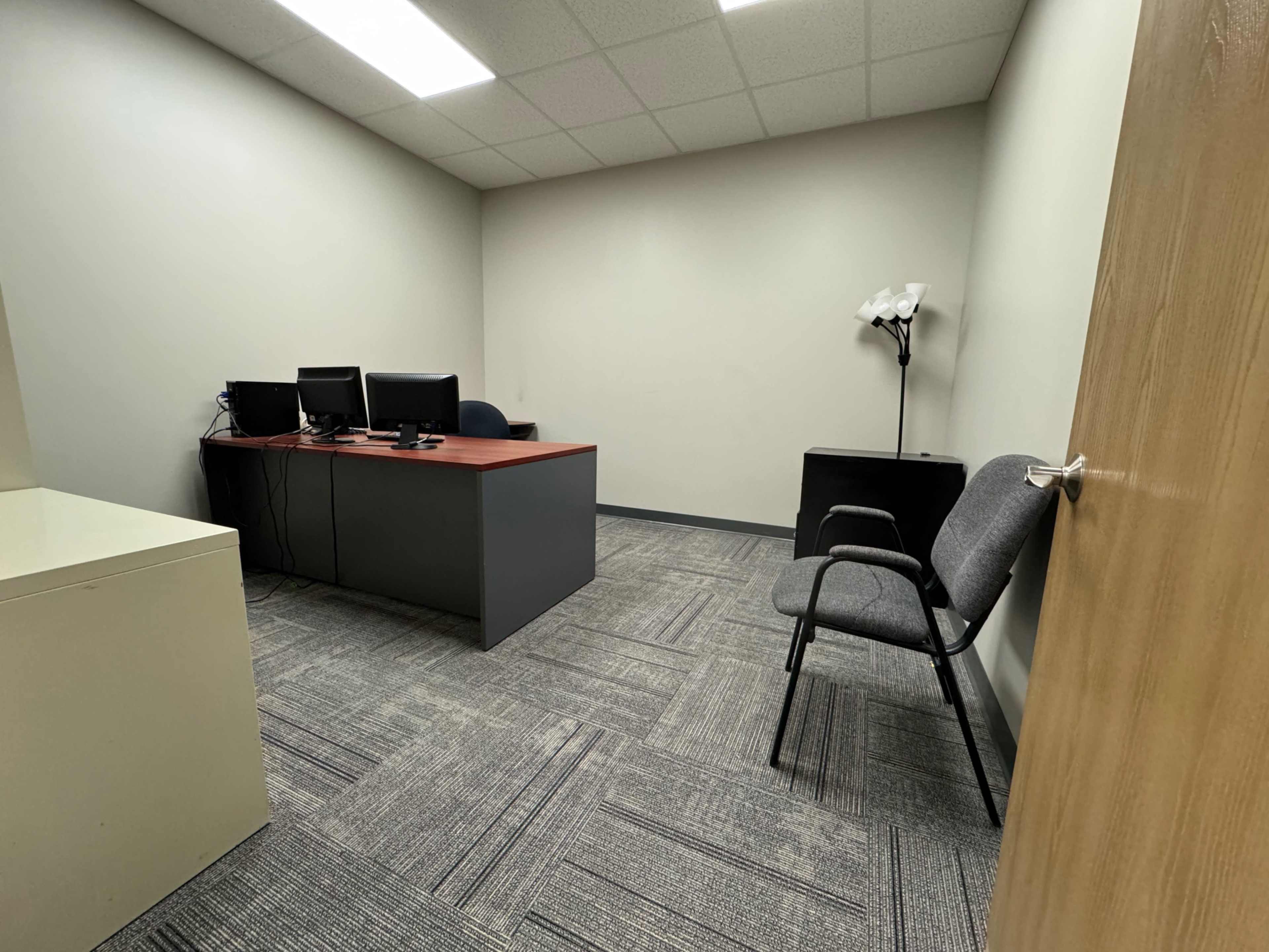 The image shows a small, empty office space with a desk, two computer monitors, a chair, and a floor lamp.