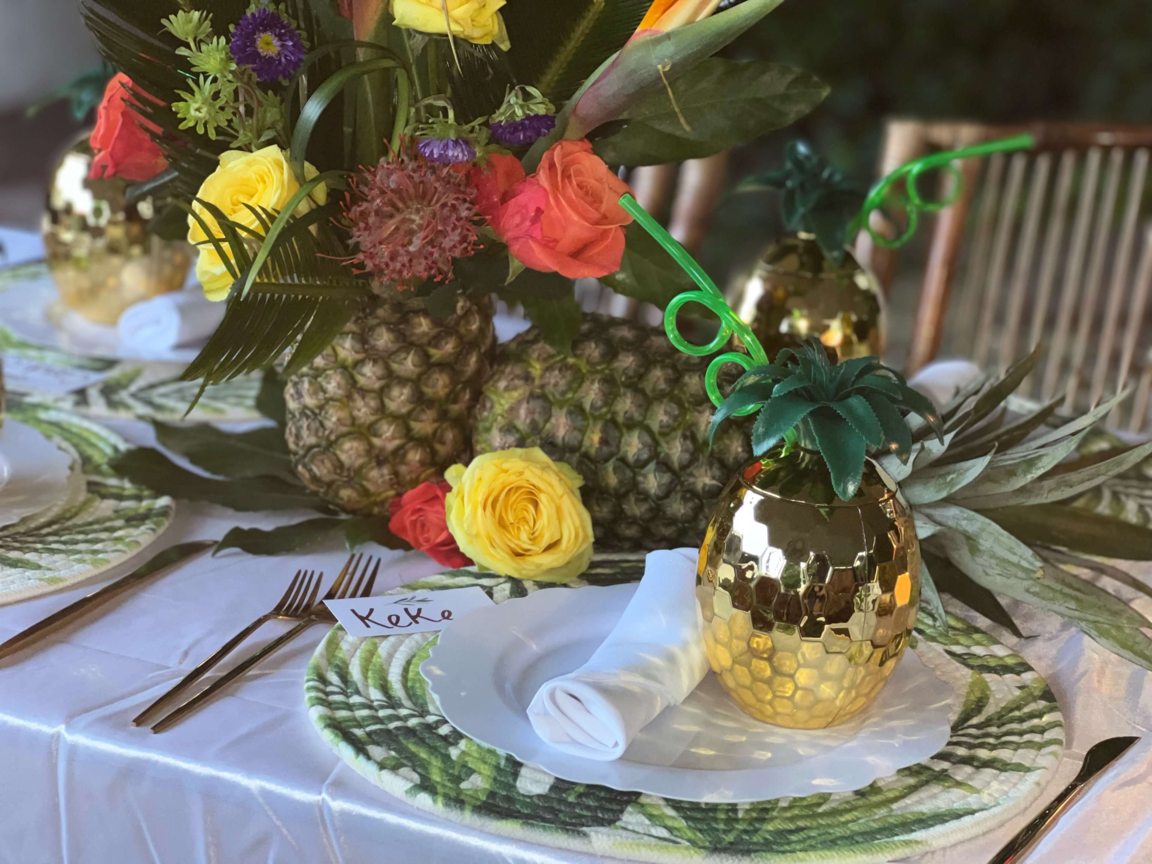A festive table setting features decorative pineapples, vibrant flowers, and gold-accented tableware arranged on a white tablecloth.