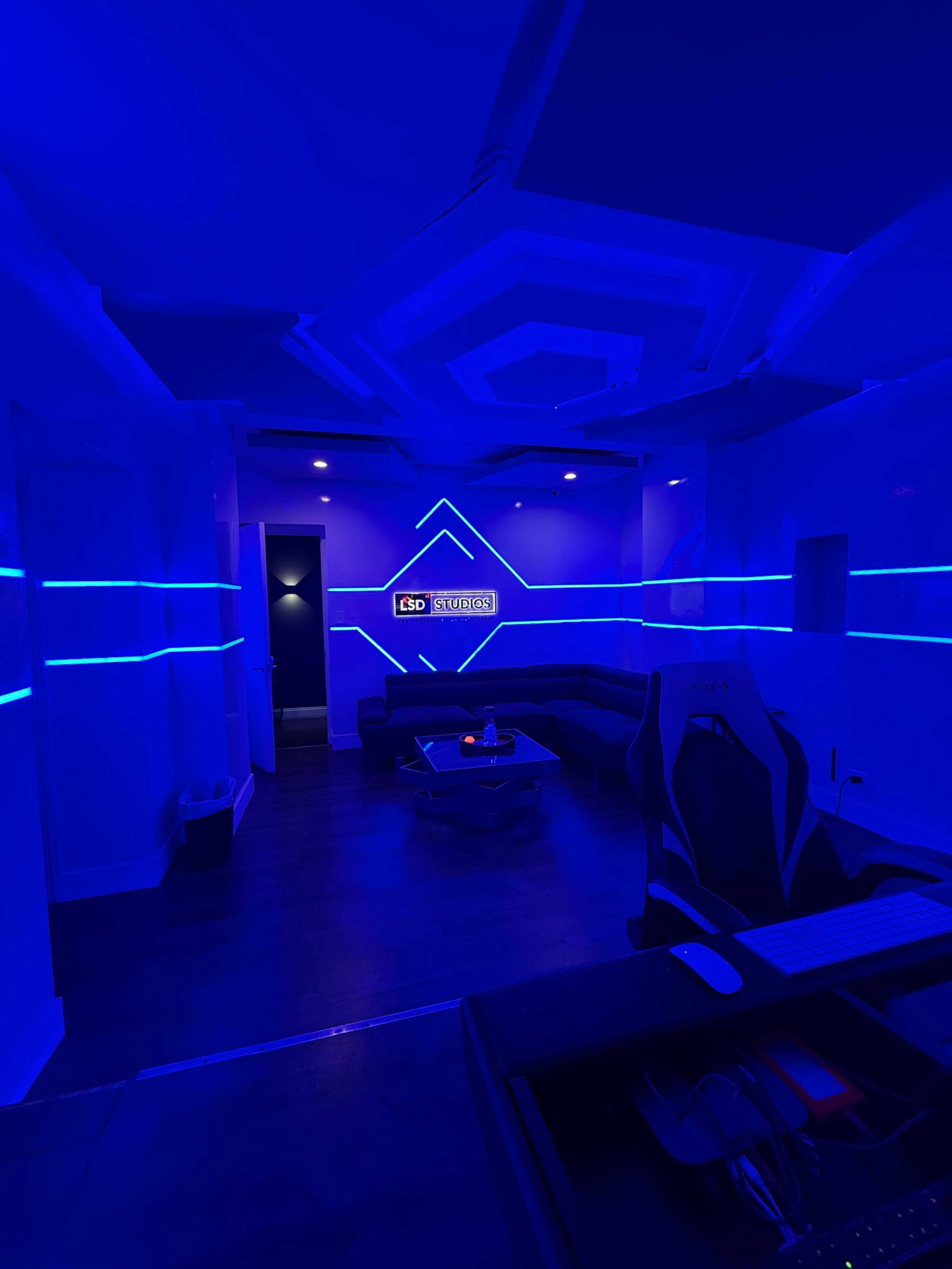 The image shows a modern room with blue neon lighting, a curved couch, and a desk with a gaming chair.
