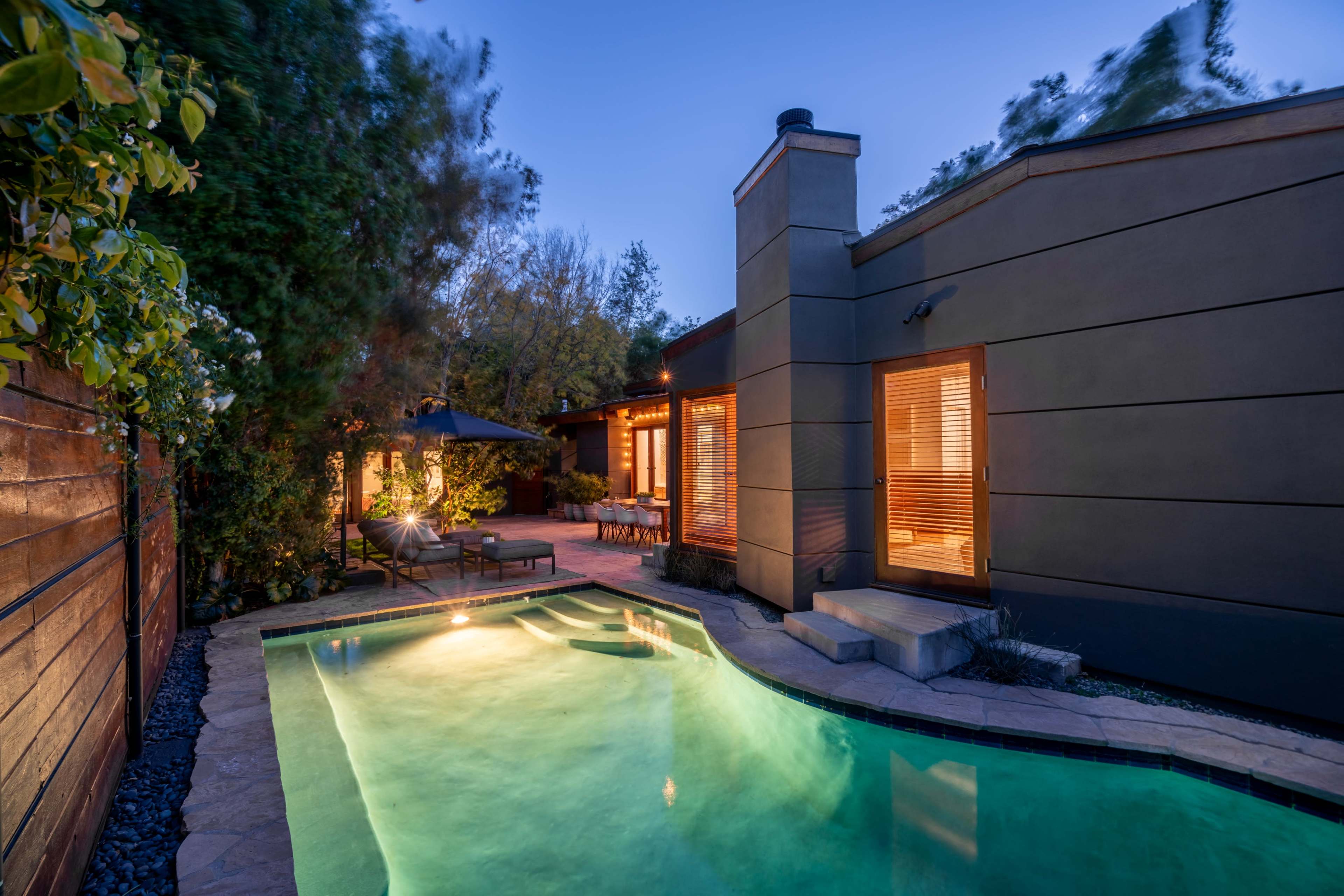 A modern backyard with a small illuminated pool, a patio with seating, and a house featuring large windows at dusk.