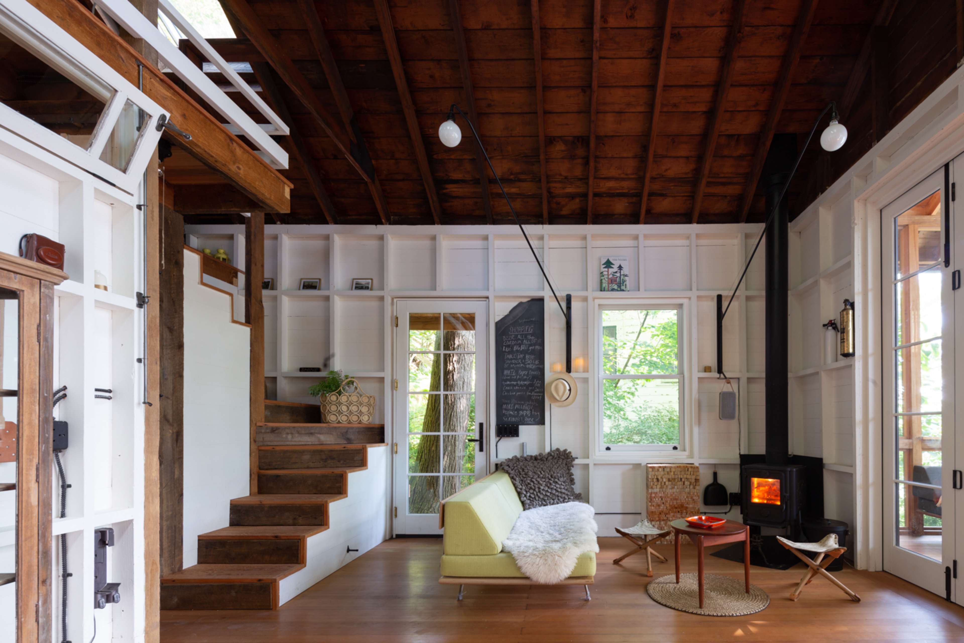The image shows a cozy interior of a cabin featuring wooden beams, a staircase, a small seating area with a white throw blanket, and a wood-burning stove.