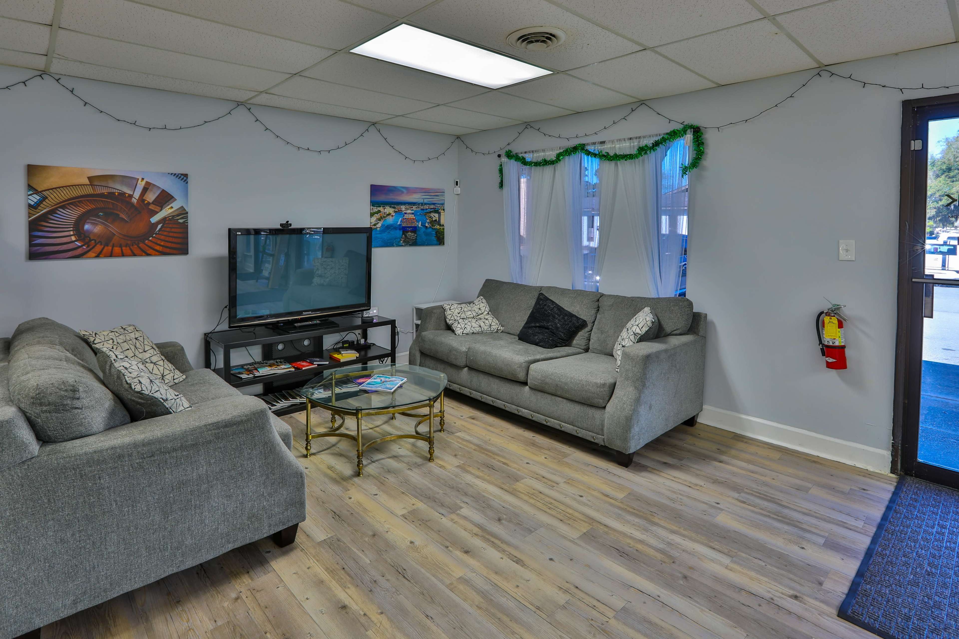 A living room with two gray couches, a television on a stand, and framed pictures on the walls, illuminated by natural light from a window.