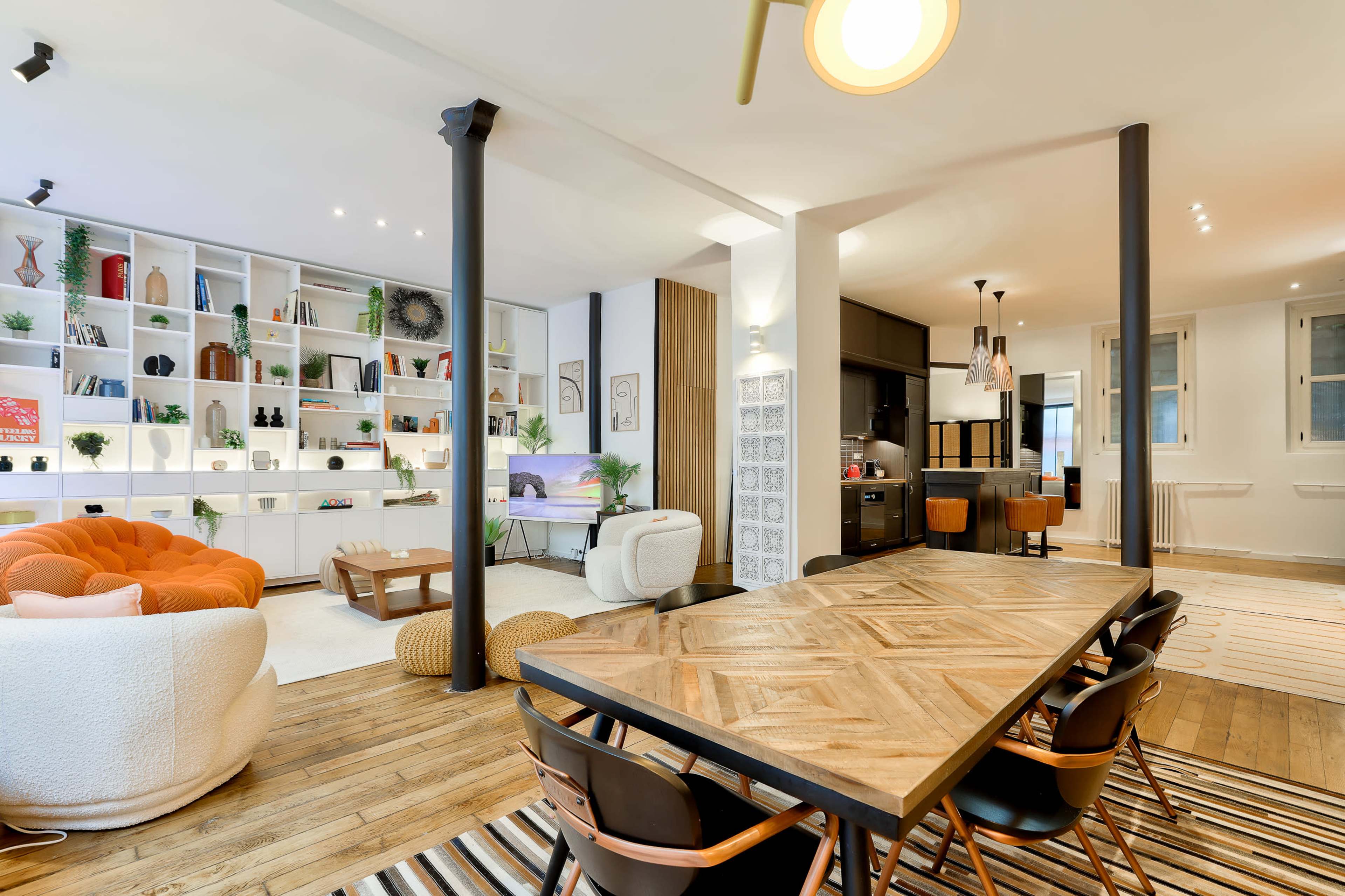 A modern living space with a dining area featuring a wooden table and upholstered chairs, alongside a bright lounge area with colorful furniture and shelves displaying decorative items.