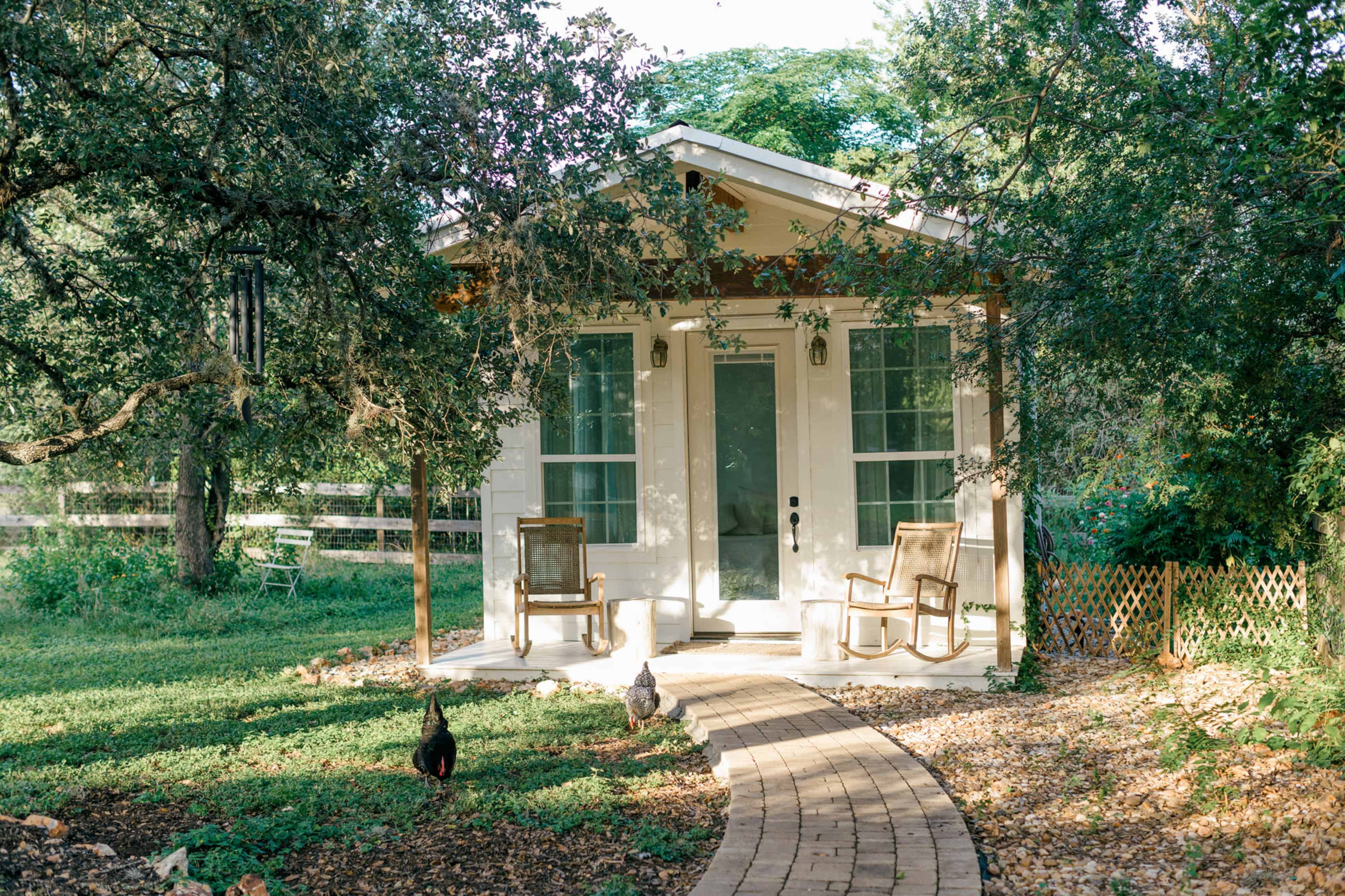 A small, white cabin with a front porch and two rocking chairs is set amidst greenery, with a winding stone pathway leading to the entrance.