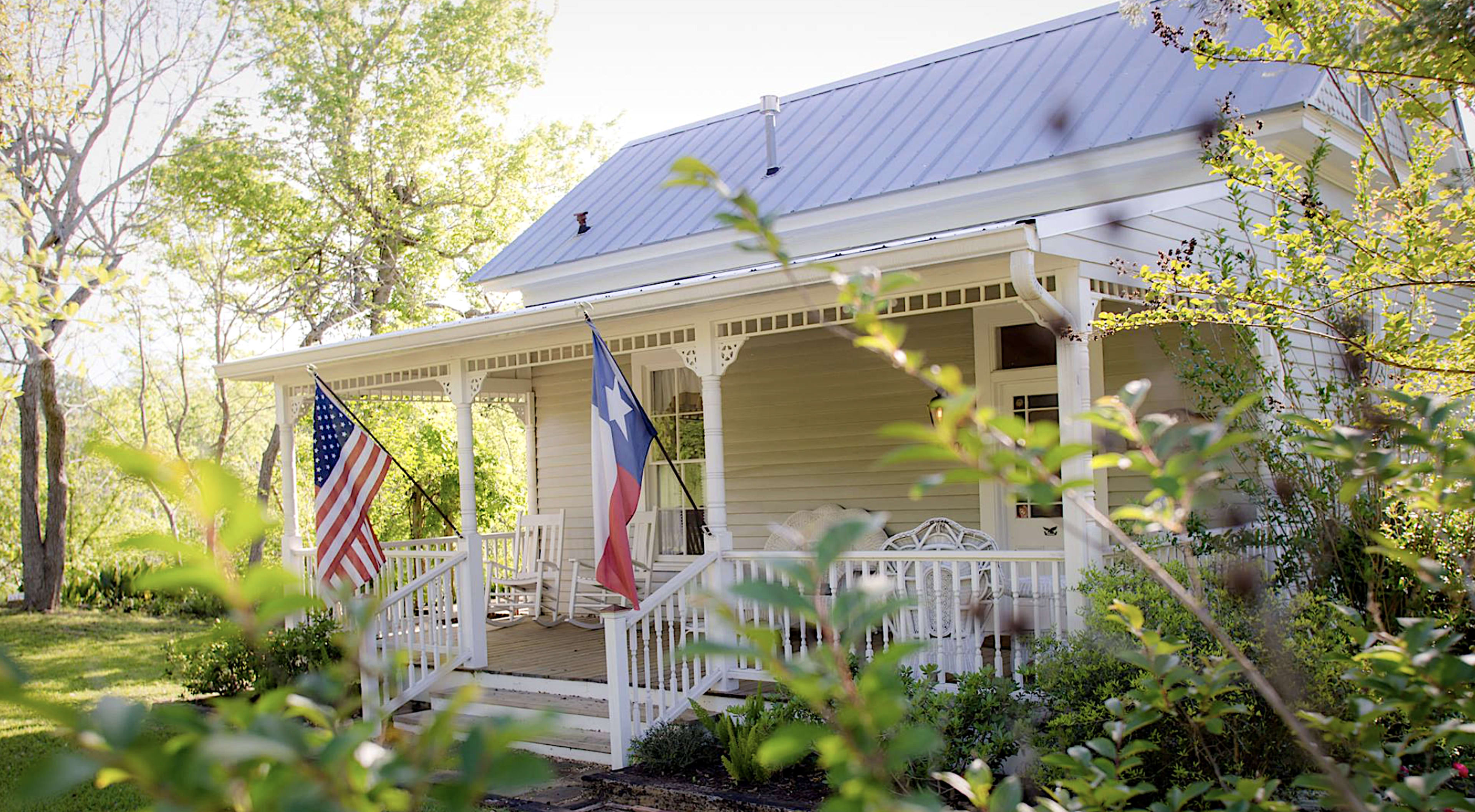A white house with a metal roof features a front porch adorned with American and Texas flags, surrounded by greenery.