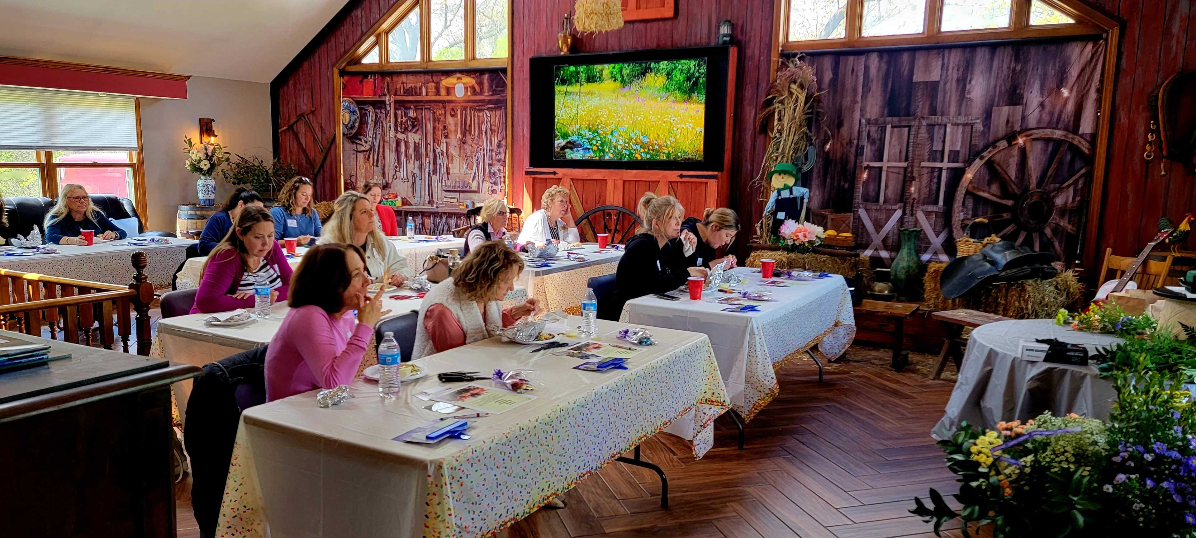A group of women sits at long tables in a rustic room decorated with farm-themed elements, engaged in a dining or workshop activity.