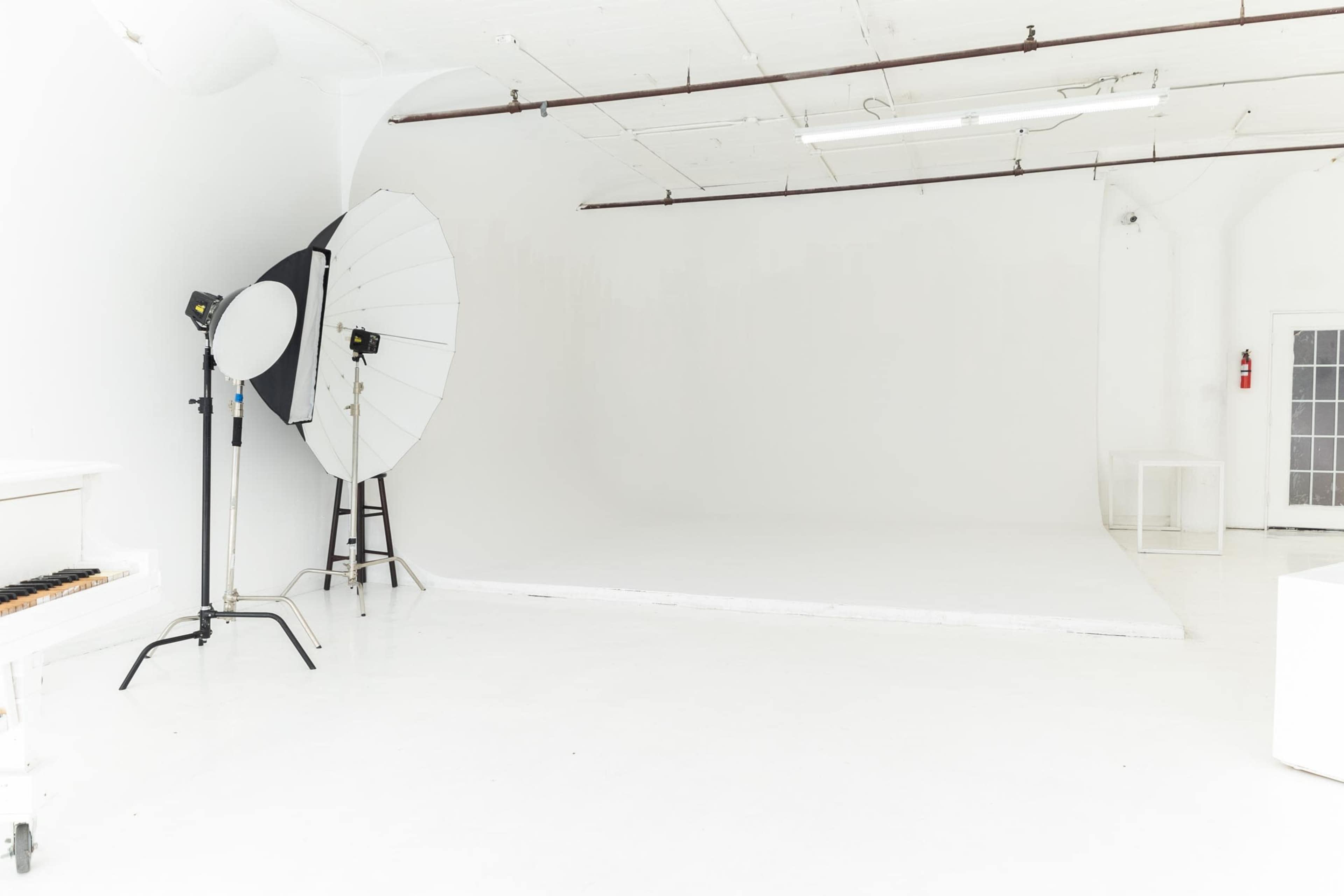 A bright, white photography studio featuring a large light umbrella and a folding chair beside a piano.