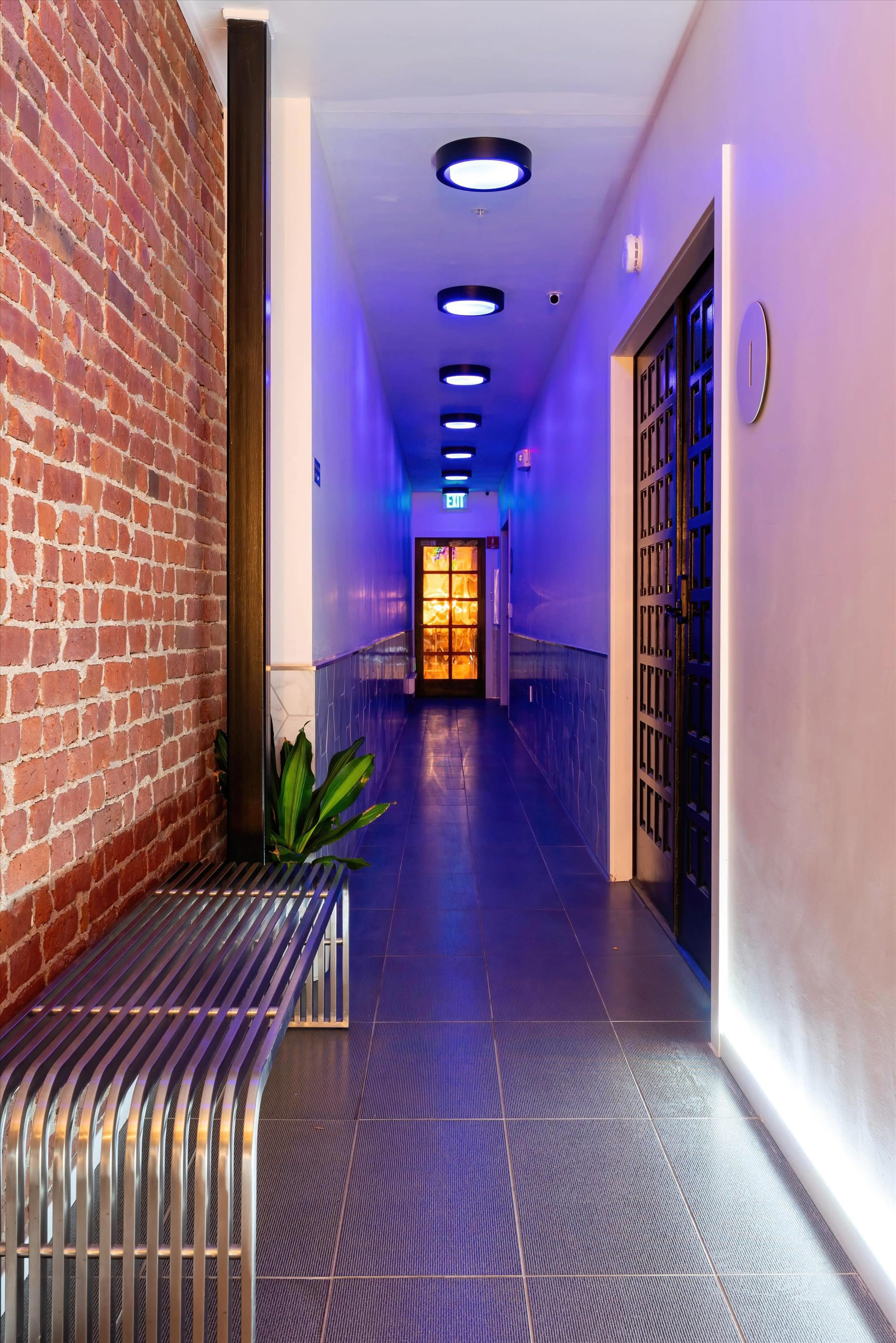 The image shows a long corridor with exposed brick walls, a metal bench, and illuminated circular ceiling lights.