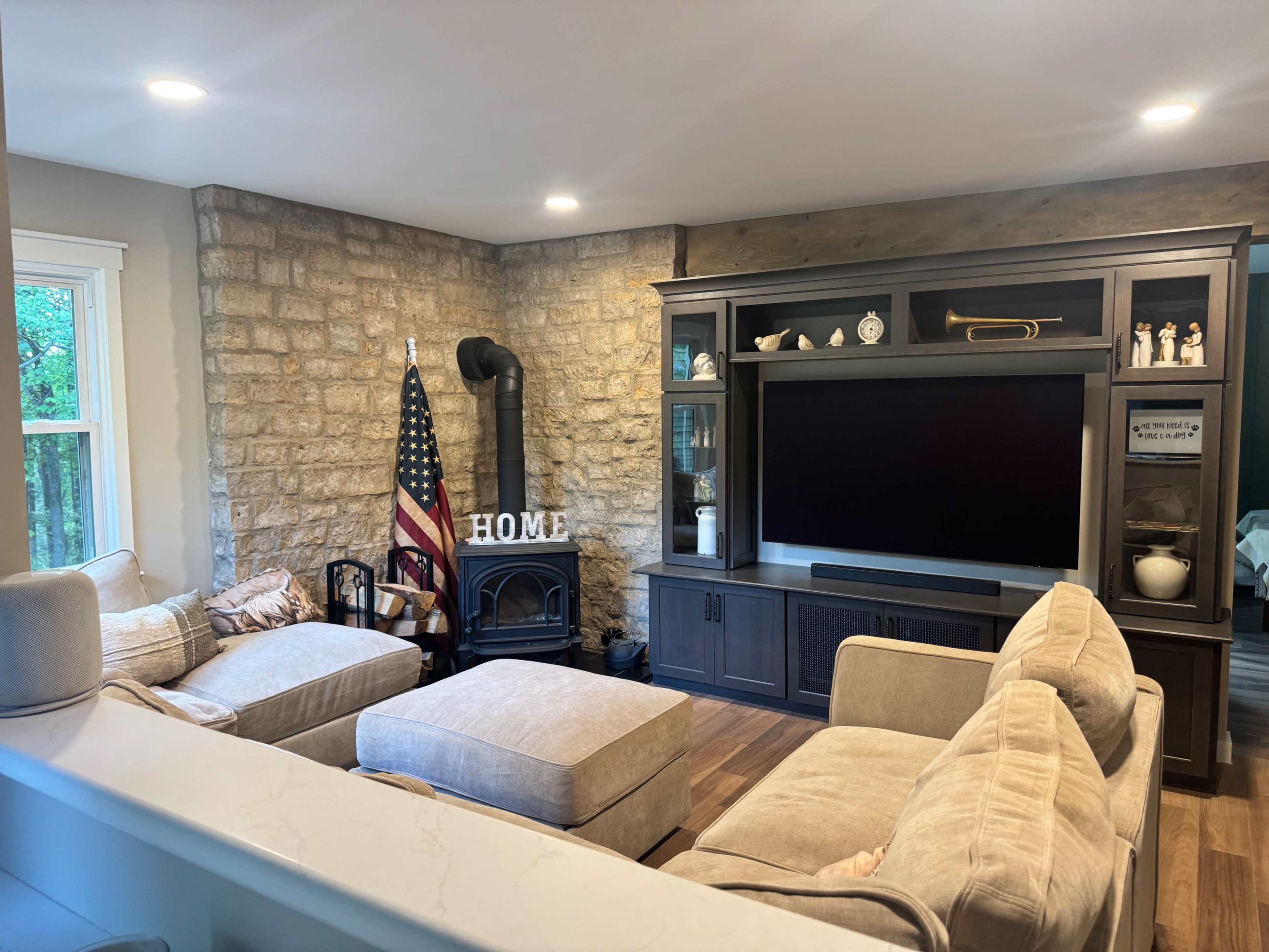 The image shows a cozy living room with a stone feature wall, a black television unit, an American flag, and a sectional sofa arranged around a dark wood floor.