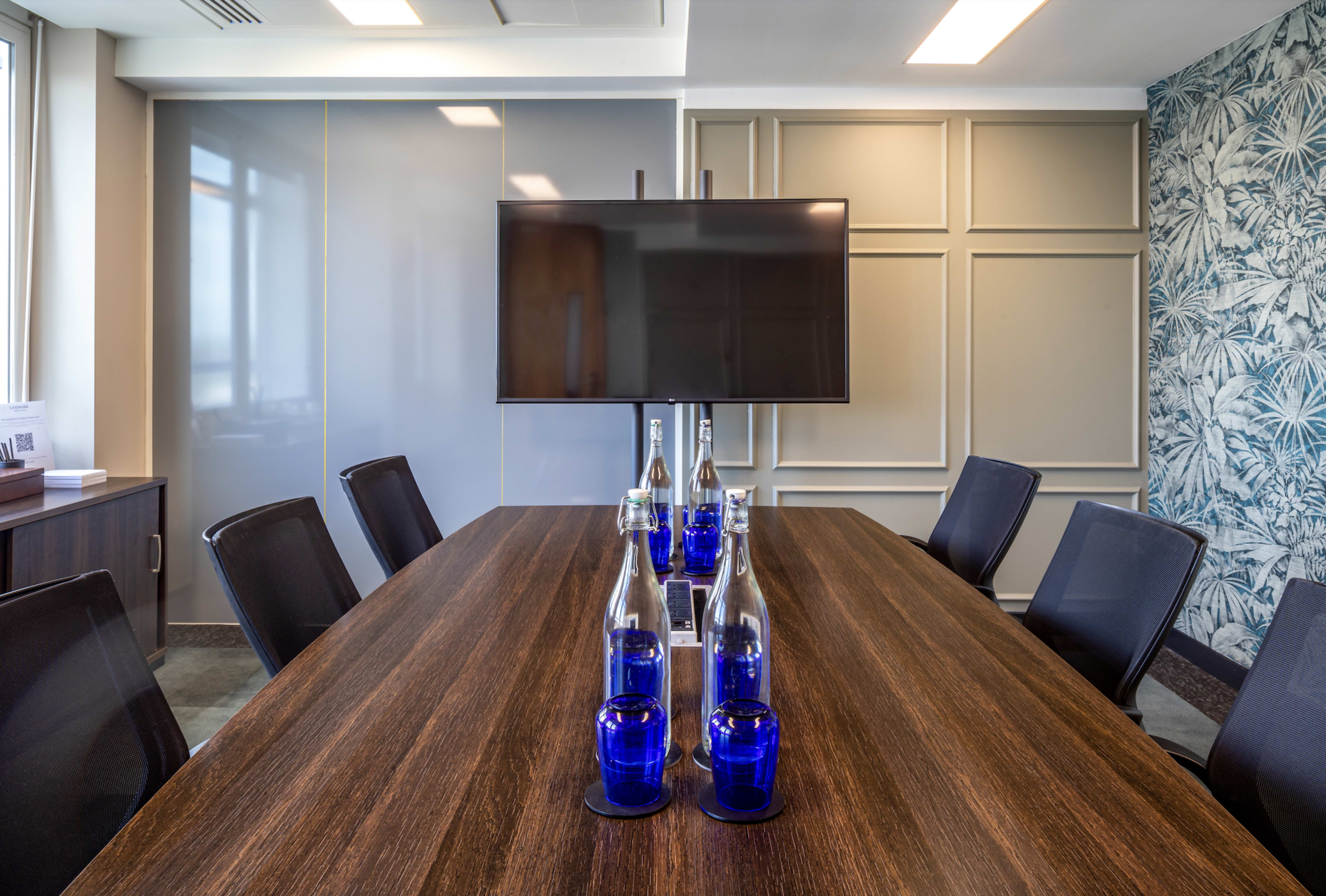 A modern conference room features a long wooden table set with glass water bottles and blue glasses, along with a wall-mounted television and patterned wallpaper.