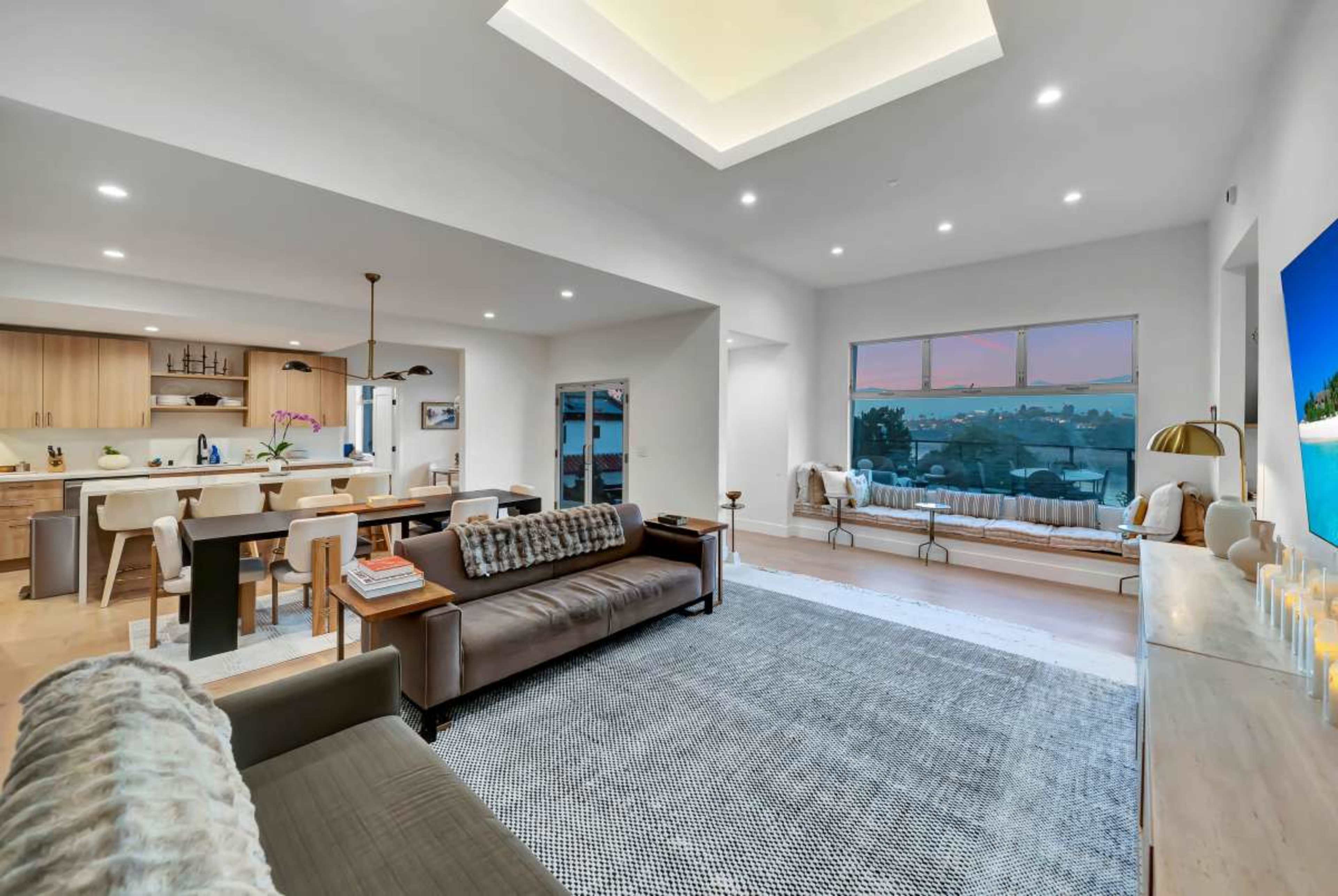 The image depicts a modern living room with a spacious open layout featuring a brown leather sofa, a light-colored dining area, and large windows offering a view of the outdoors.