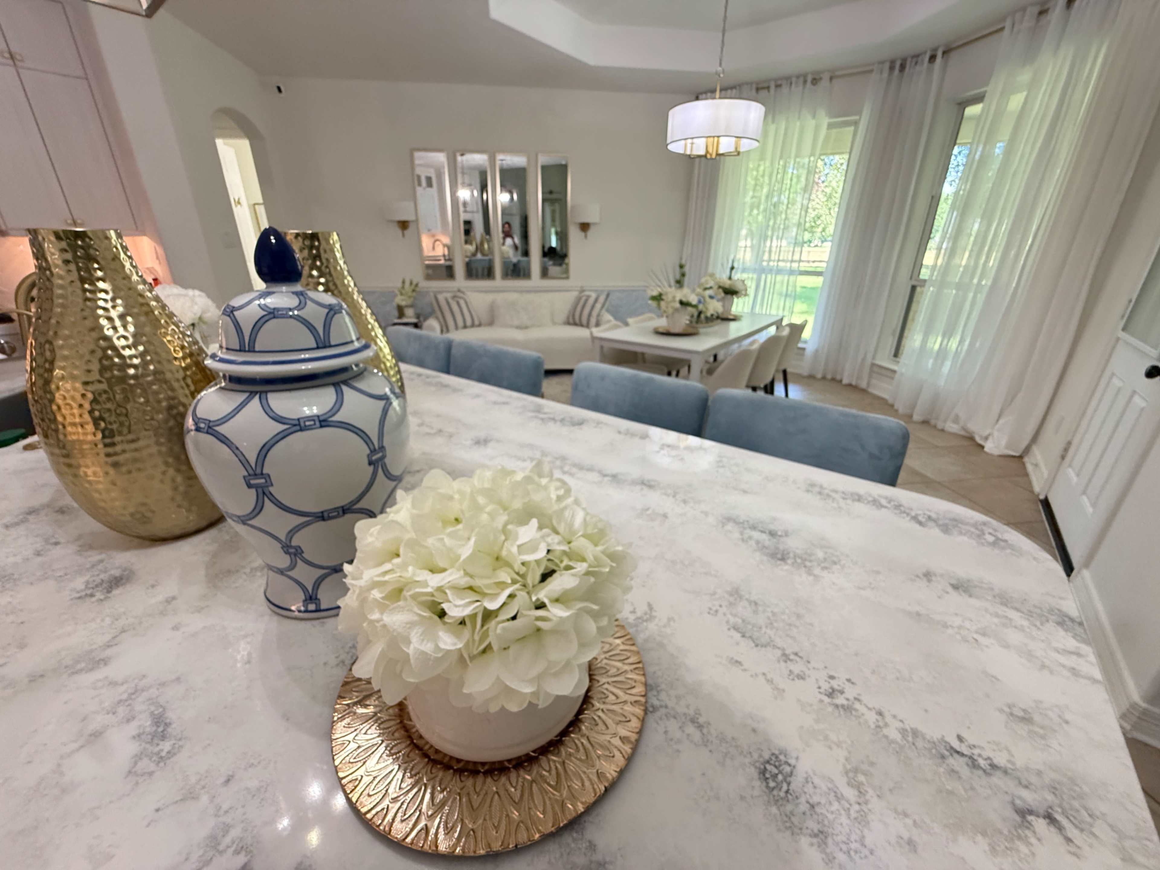 The image shows a modern dining area with a marble table, a decorative vase, and a flower arrangement, surrounded by light-colored furniture and large windows letting in natural light.