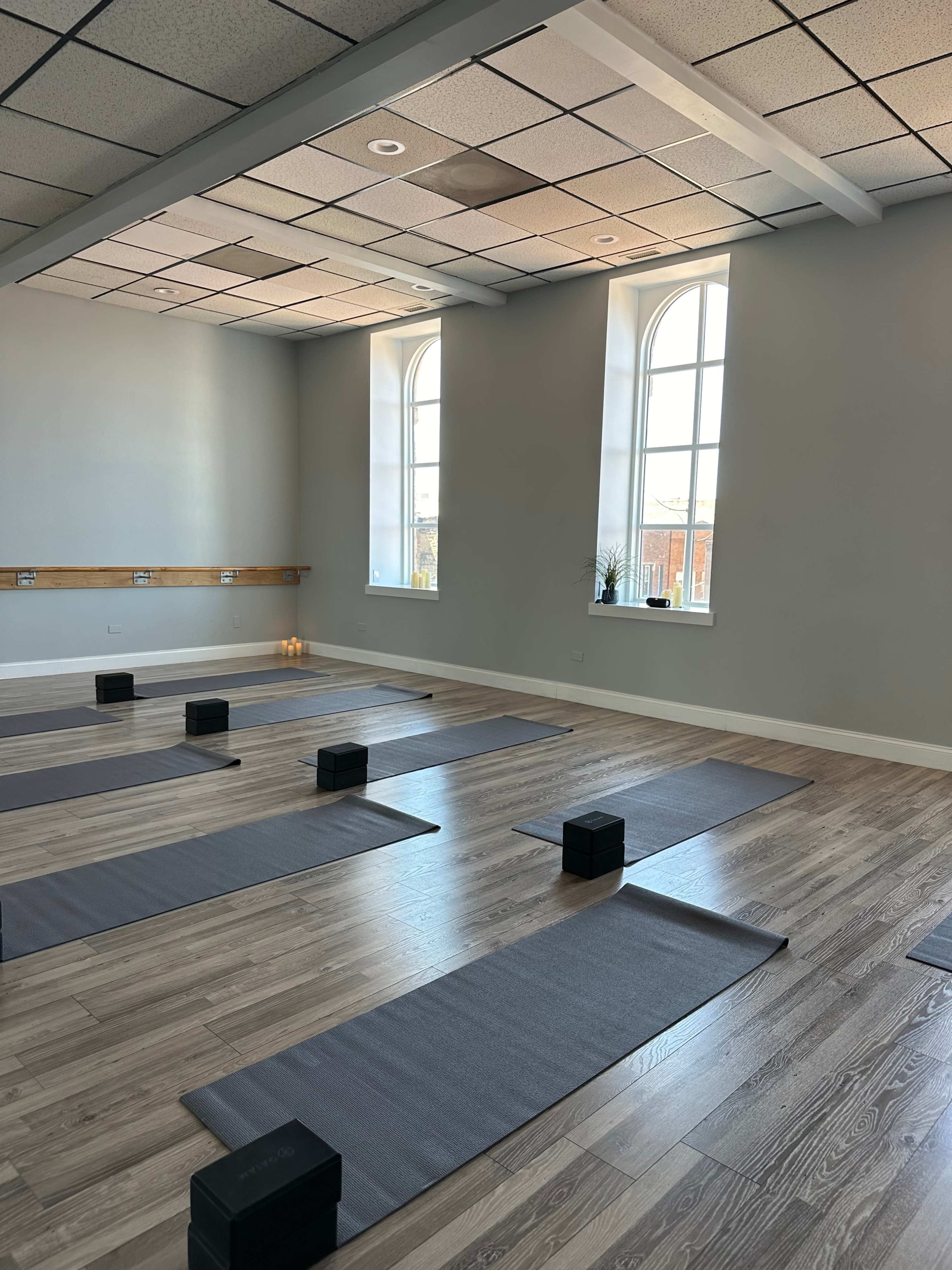 A minimalist yoga studio with several gray yoga mats arranged neatly on the wooden floor, and black blocks placed on each mat near the windows.