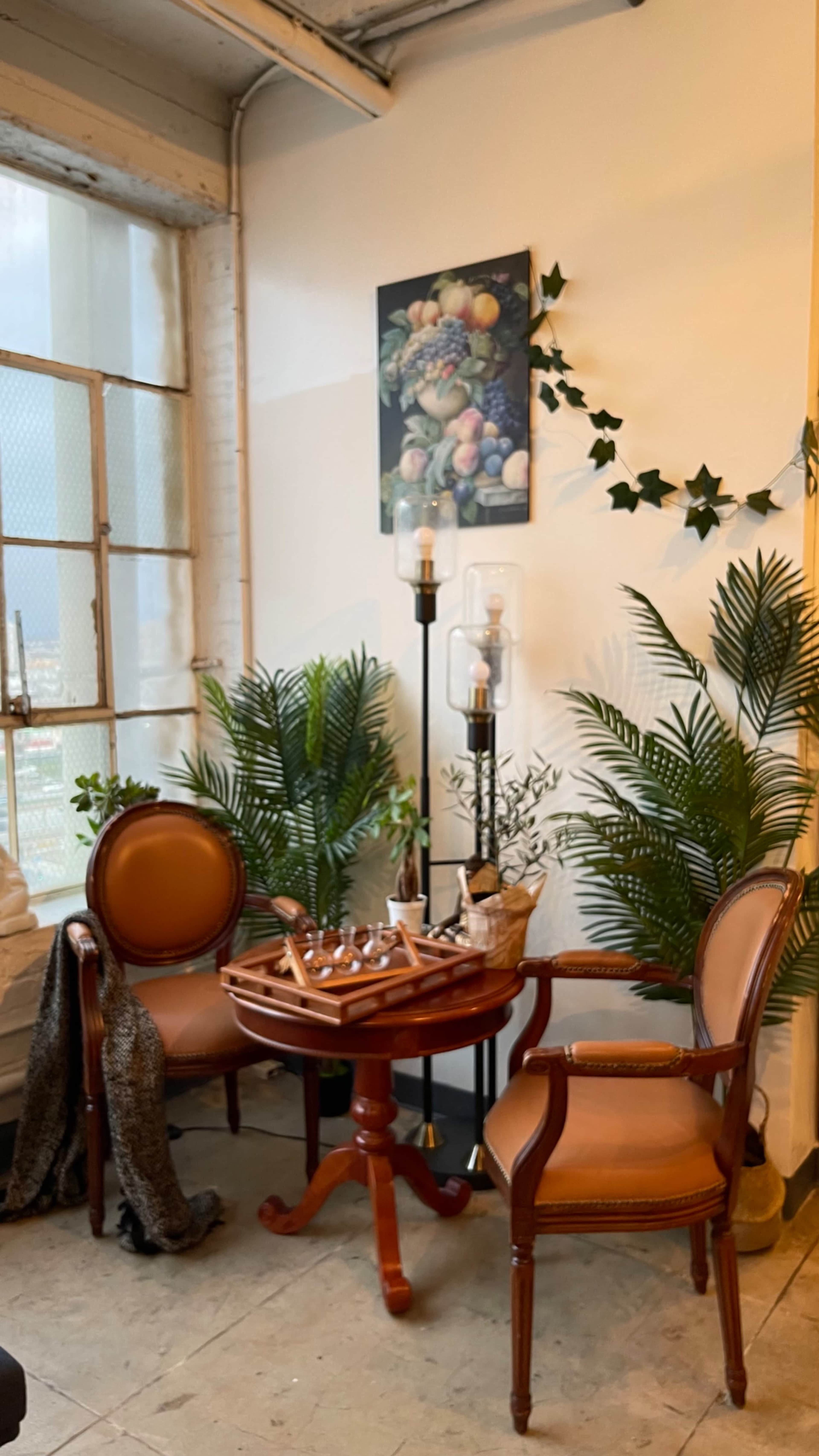 A cozy seating area features two wooden chairs with tan upholstery, a round table with various decorative items, plants, and a painting of fruit on the wall.