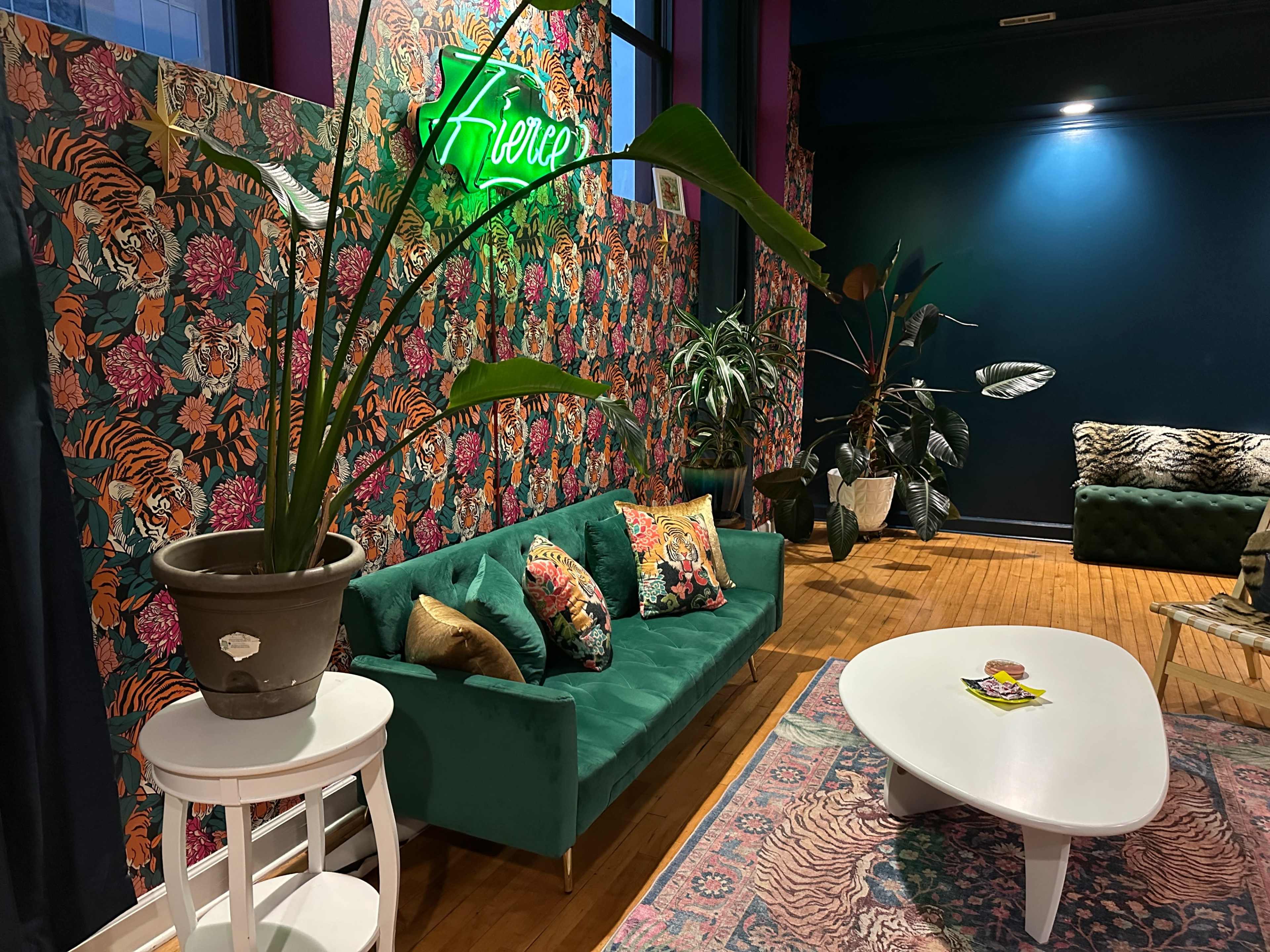 The scene features a colorful interior with a green velvet sofa, tropical floral wallpaper, and a neon sign, surrounded by various plants and decorative items.