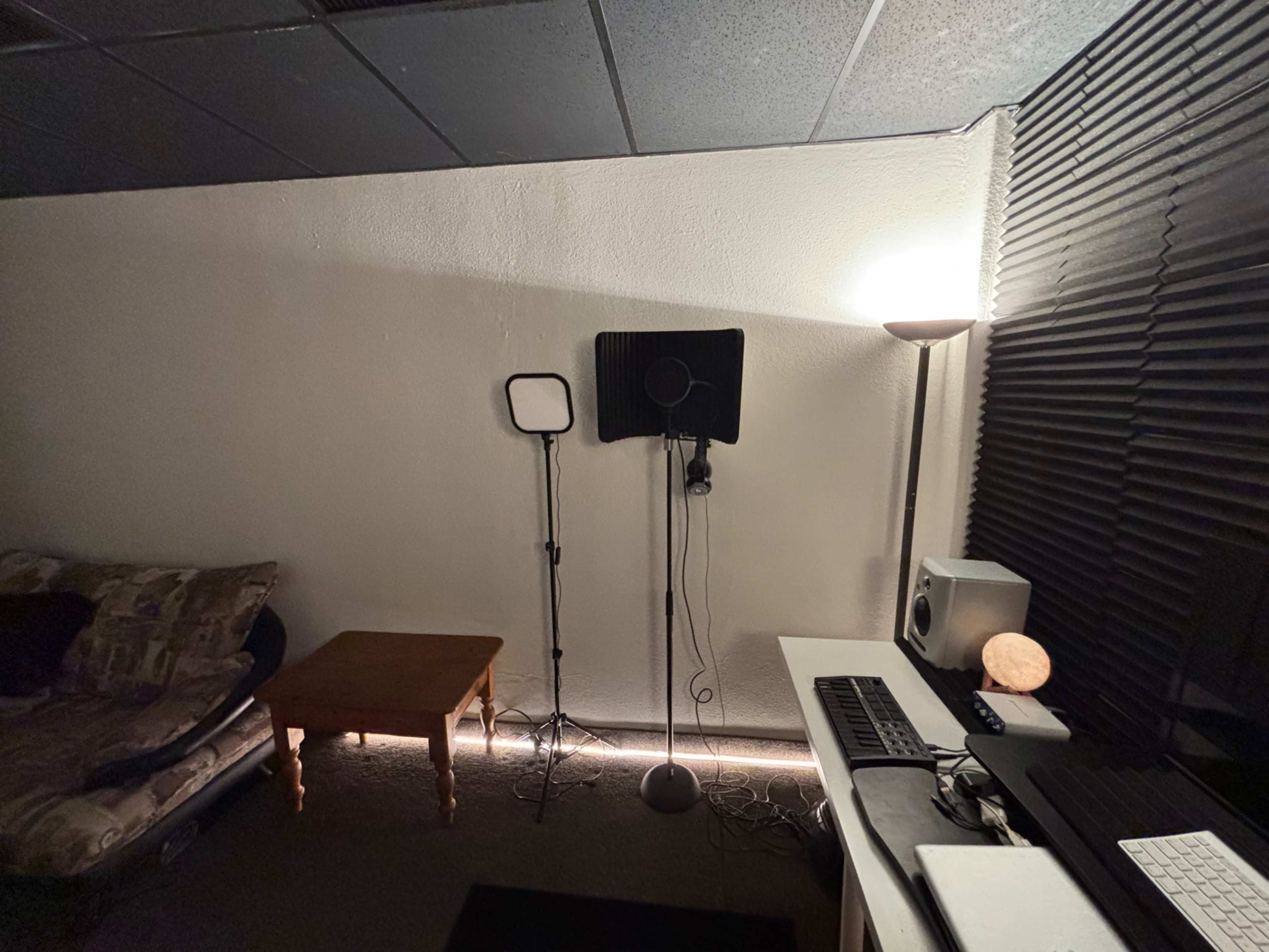 The image shows a dimly lit room with a couch, a small table, a microphone setup on stands, a lamp, and a computer workstation.