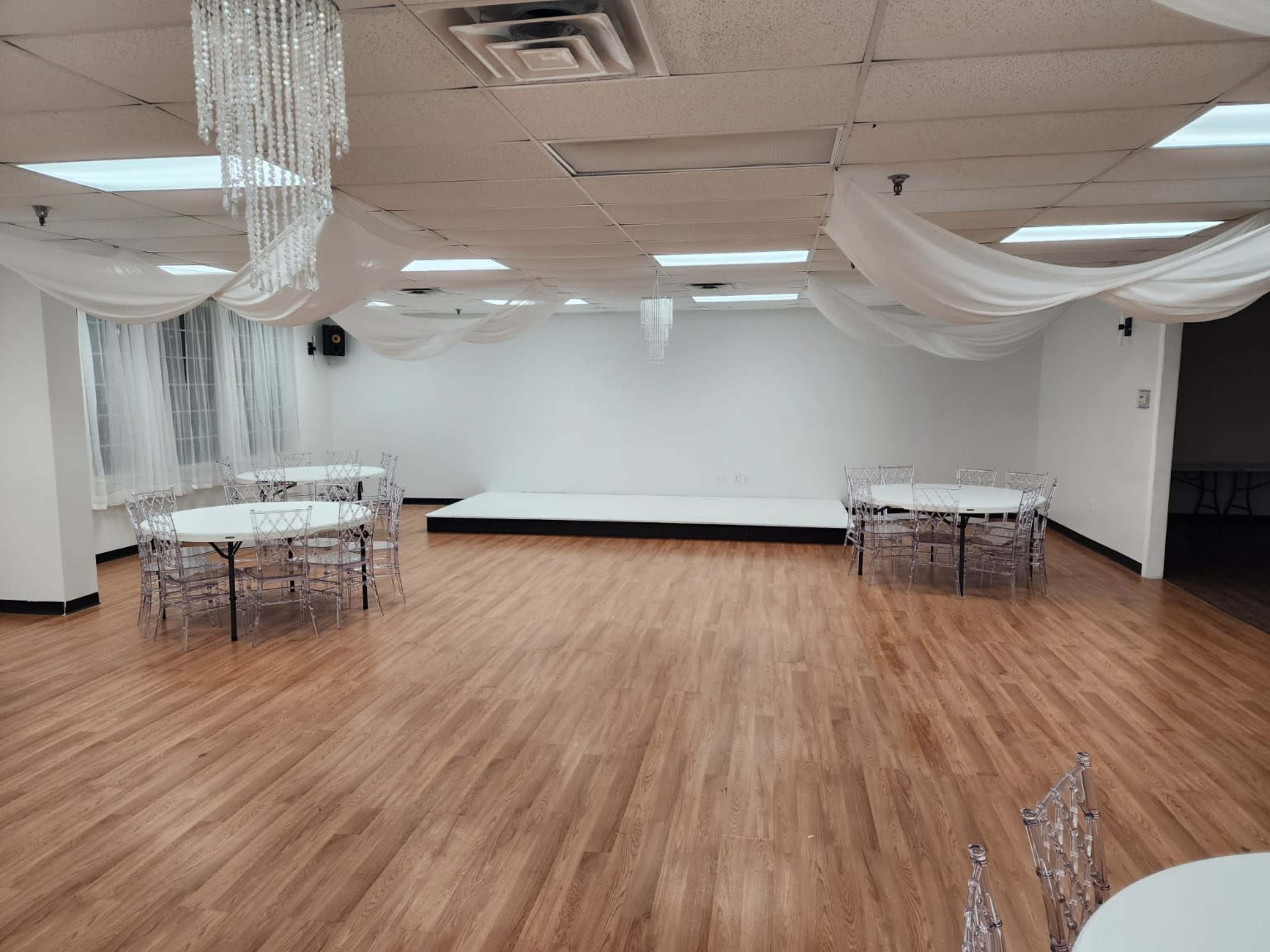 The image shows an empty event space with wooden flooring, several round tables with clear chairs, and draped fabric hanging from the ceiling.