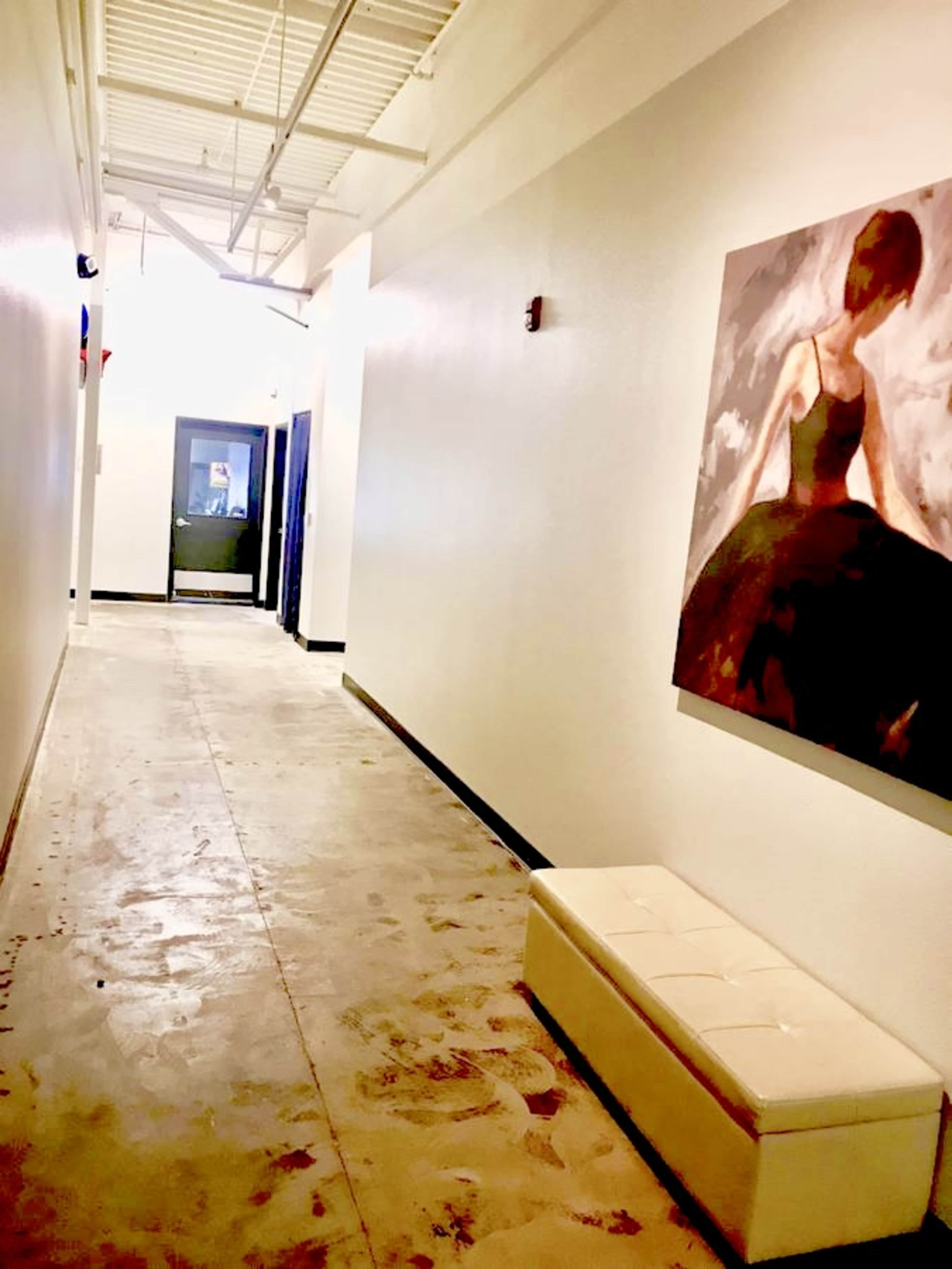 A long, white corridor features a glossy concrete floor and a large painting of a figure in a gown on one wall, with a bench positioned along the opposite side.